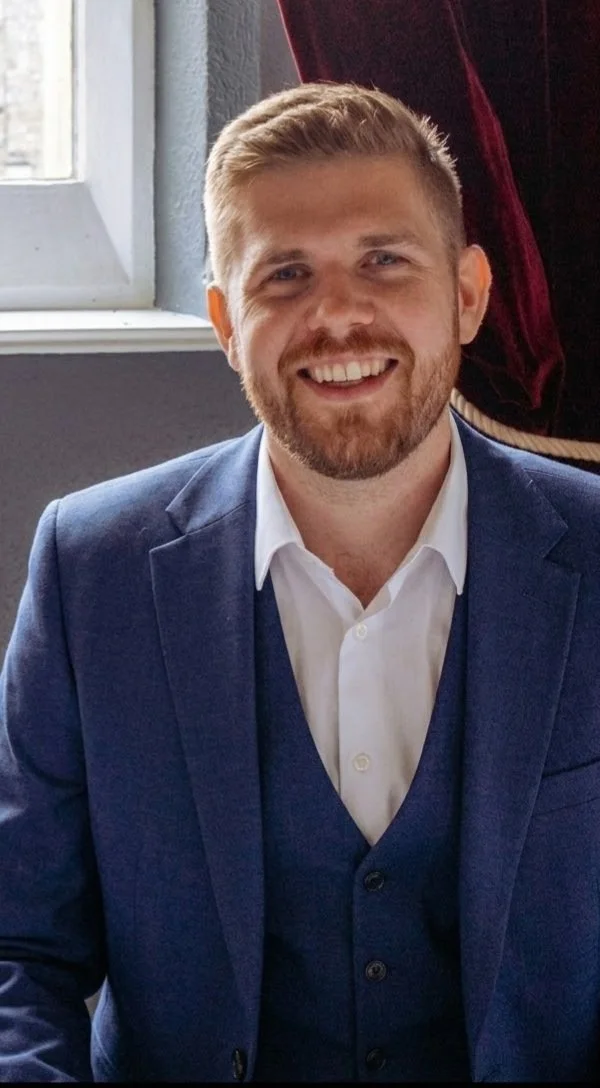 A man with blond hair and a beard smiling, dressed in a white shirt and blue blazer, sitting indoors near a window.
