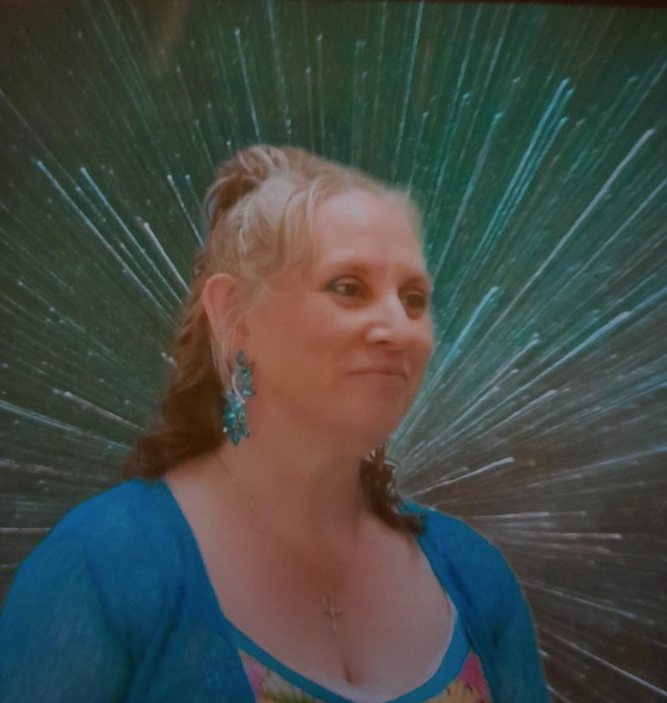 A woman with blonde hair wearing blue earrings and a blue top, standing in front of a colorful, starburst-style background.
