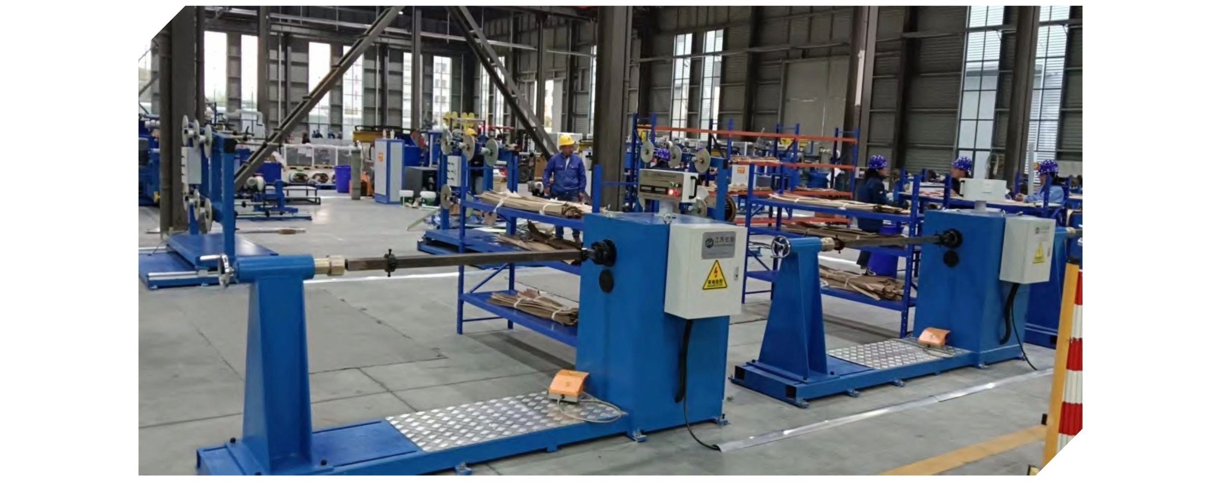 Inside a factory with blue machinery and workers wearing safety helmets working on production lines, surrounded by metal structures and large windows.