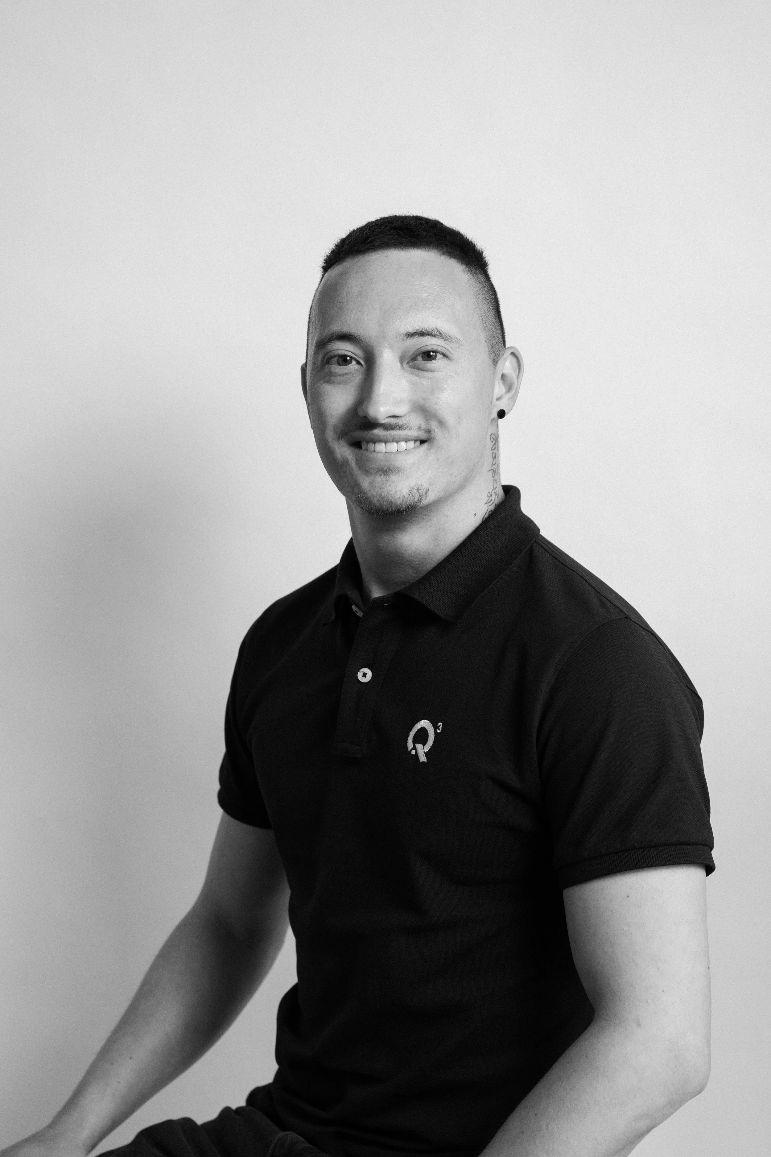 Schwarz-weiß Porträt eines Mannes mit Brille, kurzen Haaren und Bart, trägt ein schwarzes Polo-Shirt mit Q3-Logo, sitzt vor weißem Hintergrund und lächelt.