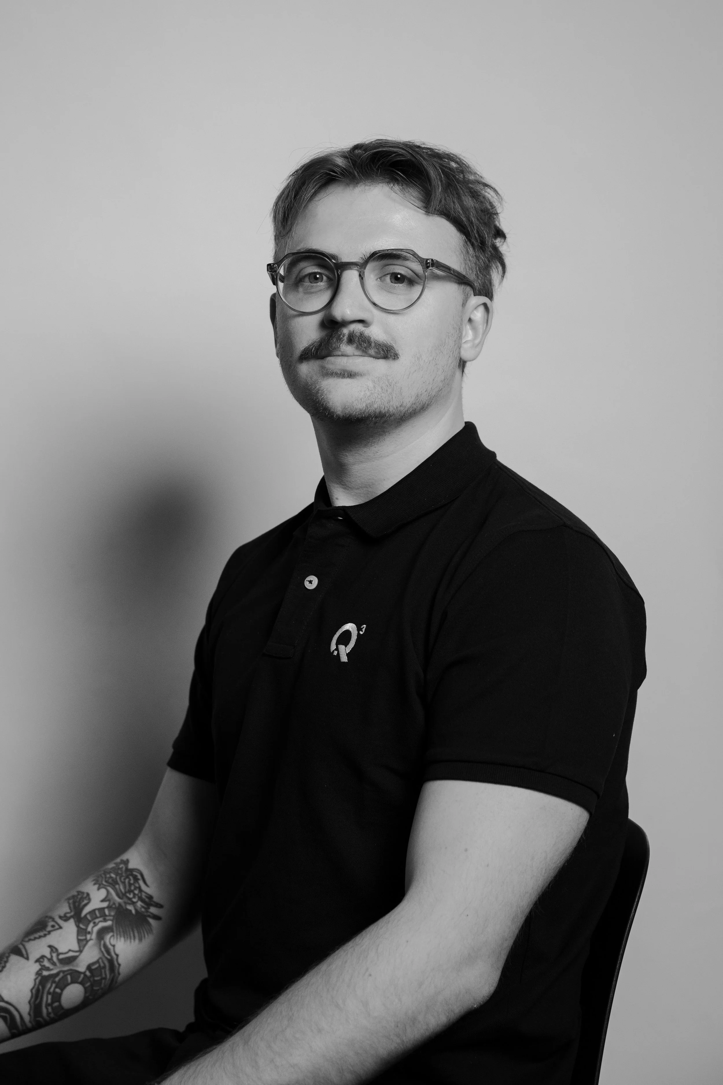 Schwarz-weiß Porträt eines Mannes mit Brille, kurzen Haaren und Bart, trägt ein schwarzes Polo-Shirt mit Q3-Logo, sitzt vor weißem Hintergrund und lächelt.
