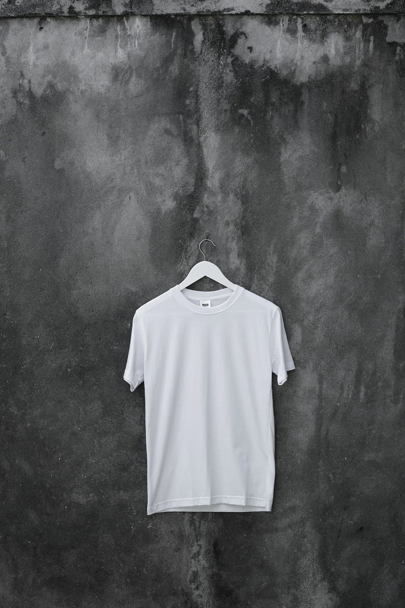 White T-shirt hanging on a white hanger against a dark, textured concrete wall.