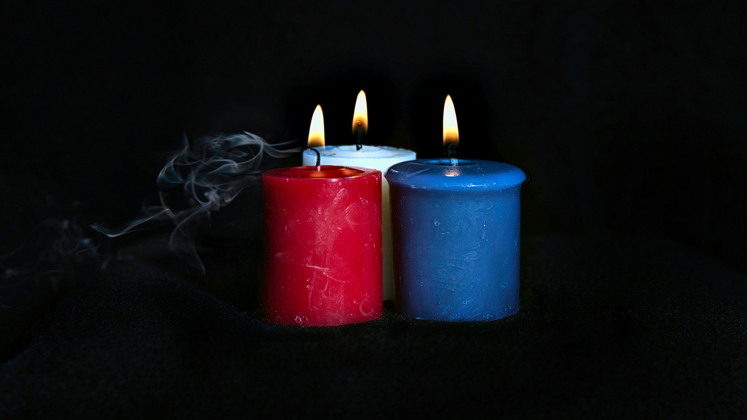 Three burning candles, red, white, and blue, on a black background with smoke rising from the red candle.