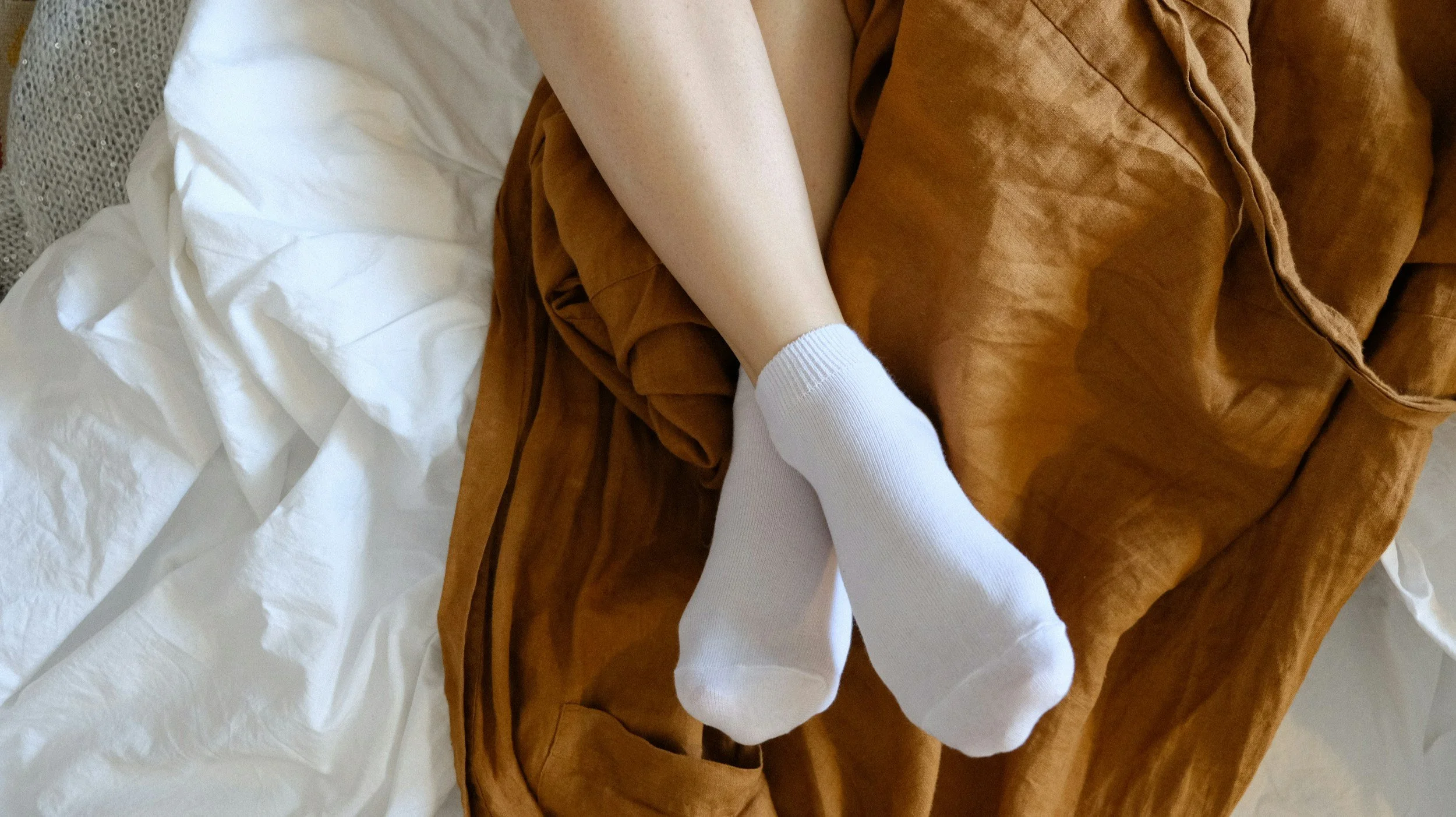 A person's legs and feet, with the feet wearing white socks, resting on a brown leather blanket or cushion on top of a white bedspread.