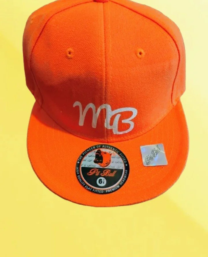 Orange baseball cap with the letters 'MB' embroidered on the front in white, with a sticker on the brim featuring a lion logo and the text 'Dri-Pull'.