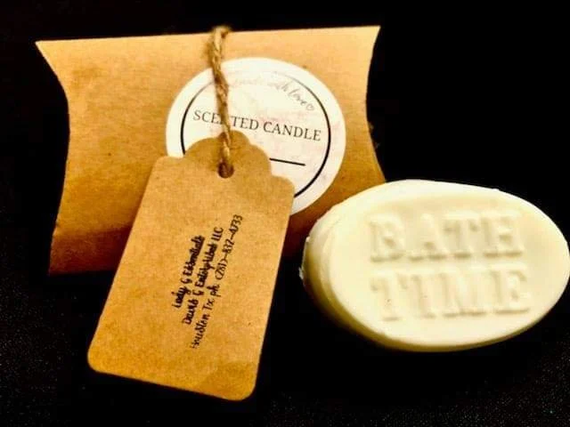 A white scented candle with the inscription 'Dreams Take Time' on it, a brown paper tag with handwritten details, and a blank rectangular label in the background.