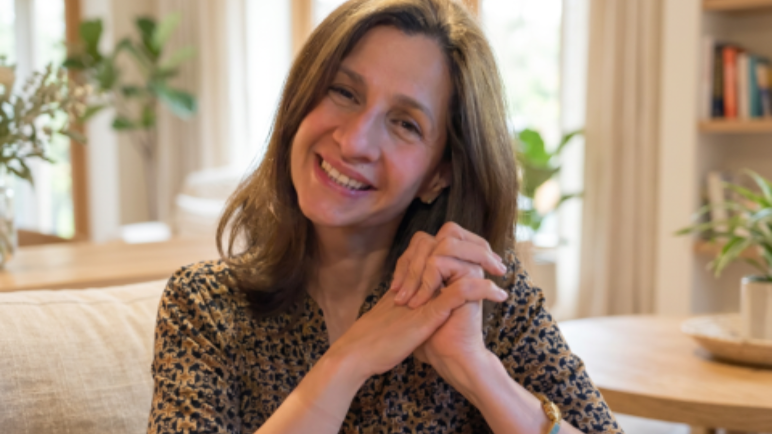 A woman with shoulder-length brown hair smiling and sitting at a table in a cozy, well-lit room with plants and books in the background.