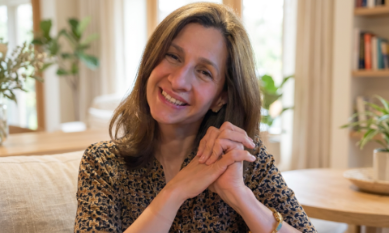 A woman with brown hair smiling and resting her hands on a table in a well-lit room with plants and bookshelves