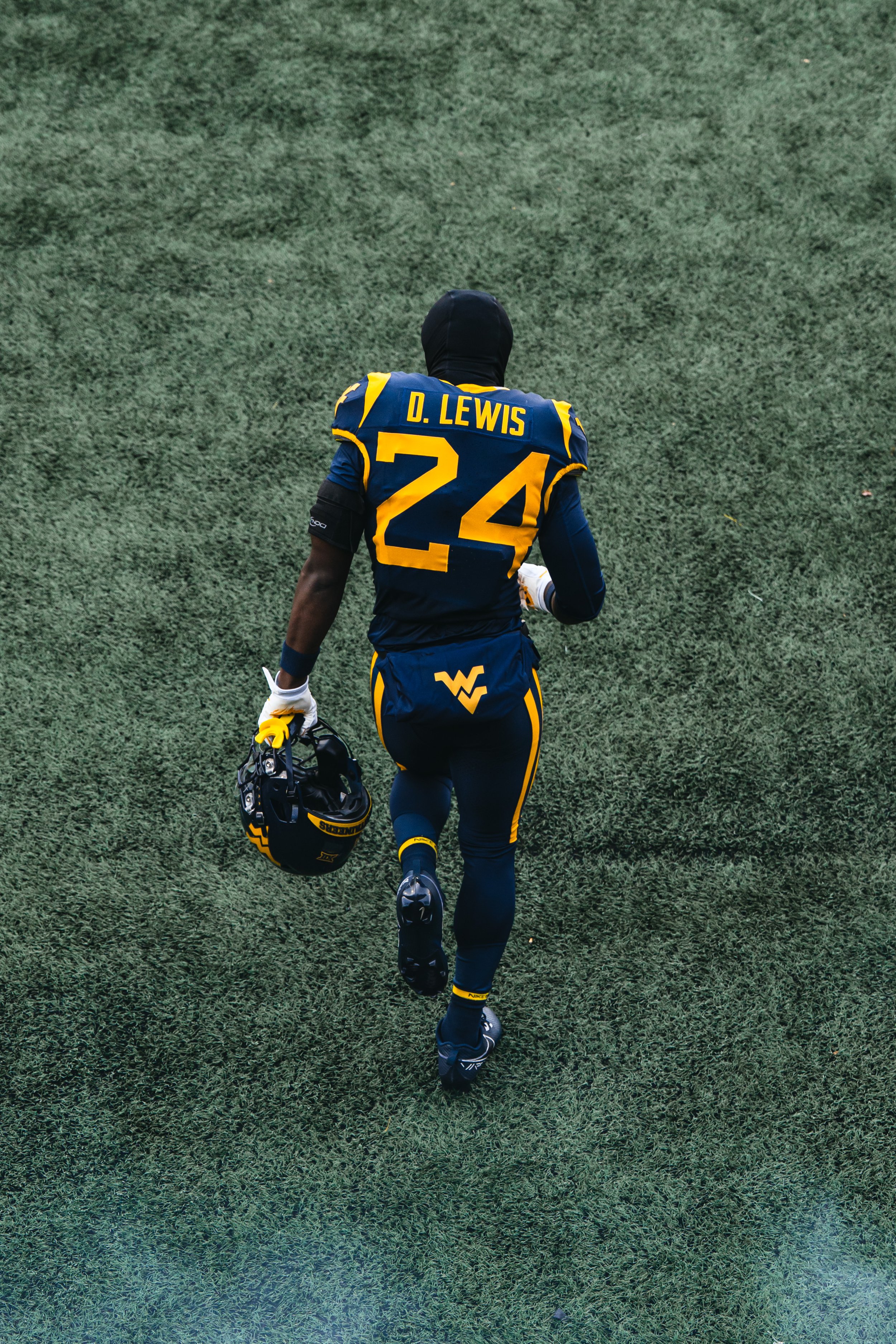 A football player from West Virginia University running on a grassy field, holding a helmet in one hand, wearing a navy blue and gold uniform with the name 'D. Lewis' and the number 24 on the back.