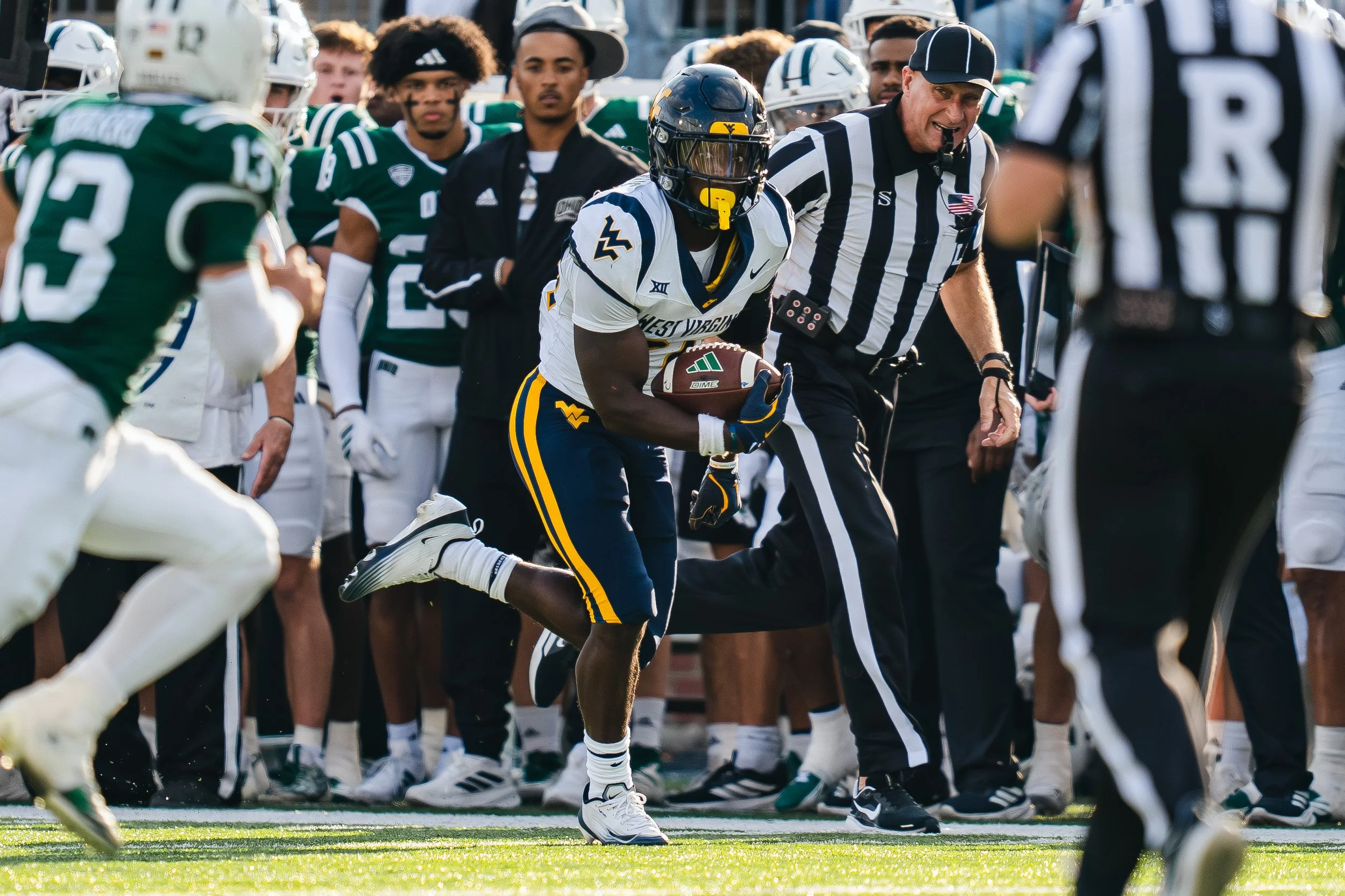 College football game with West Virginia player running with the ball, surrounded by players from opposing team and referees, with spectators in the background.