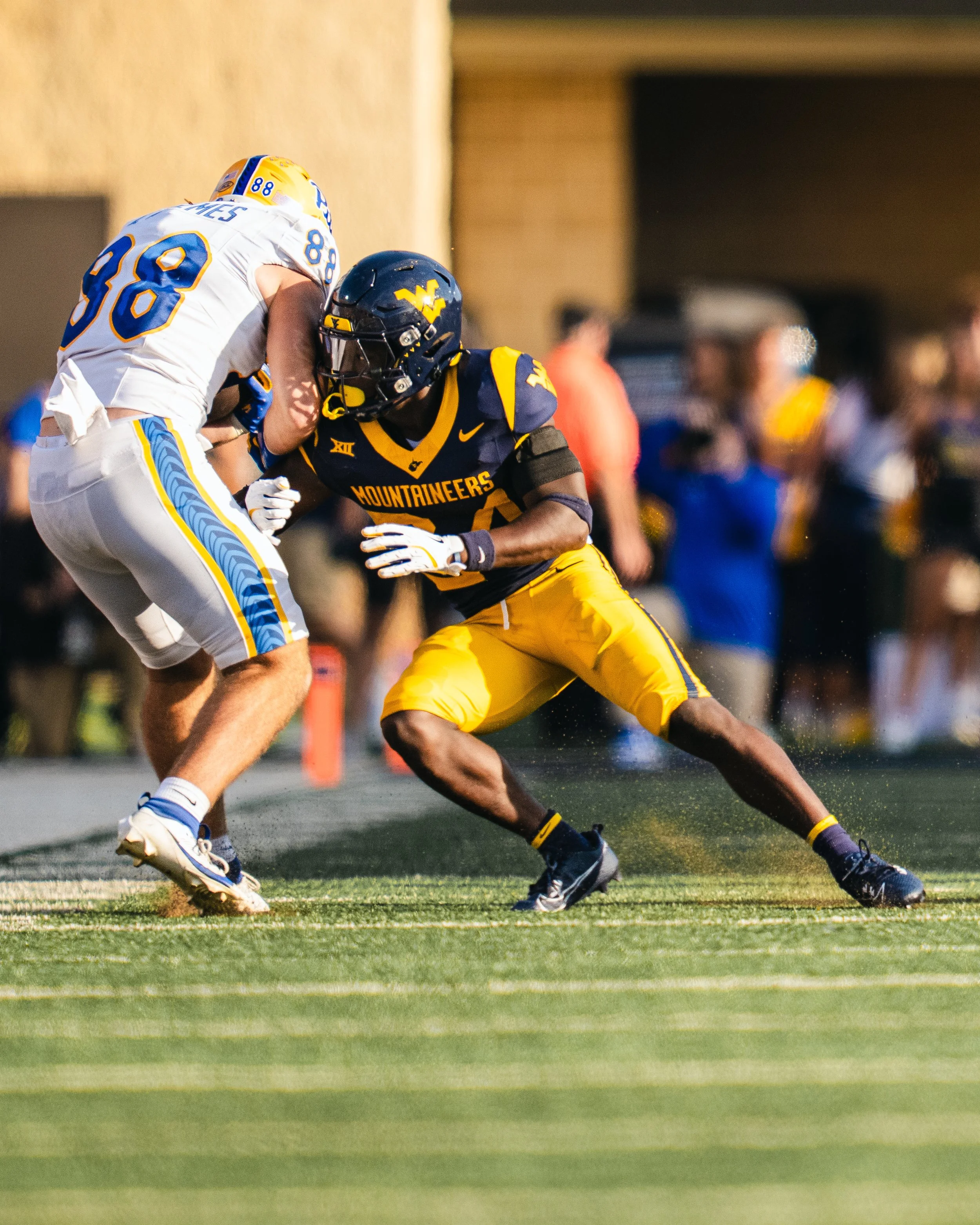 Two football players in a game, one from West Virginia Mountaineers and one from UCLA Bruins, engaged in a tackle on the field during a game.