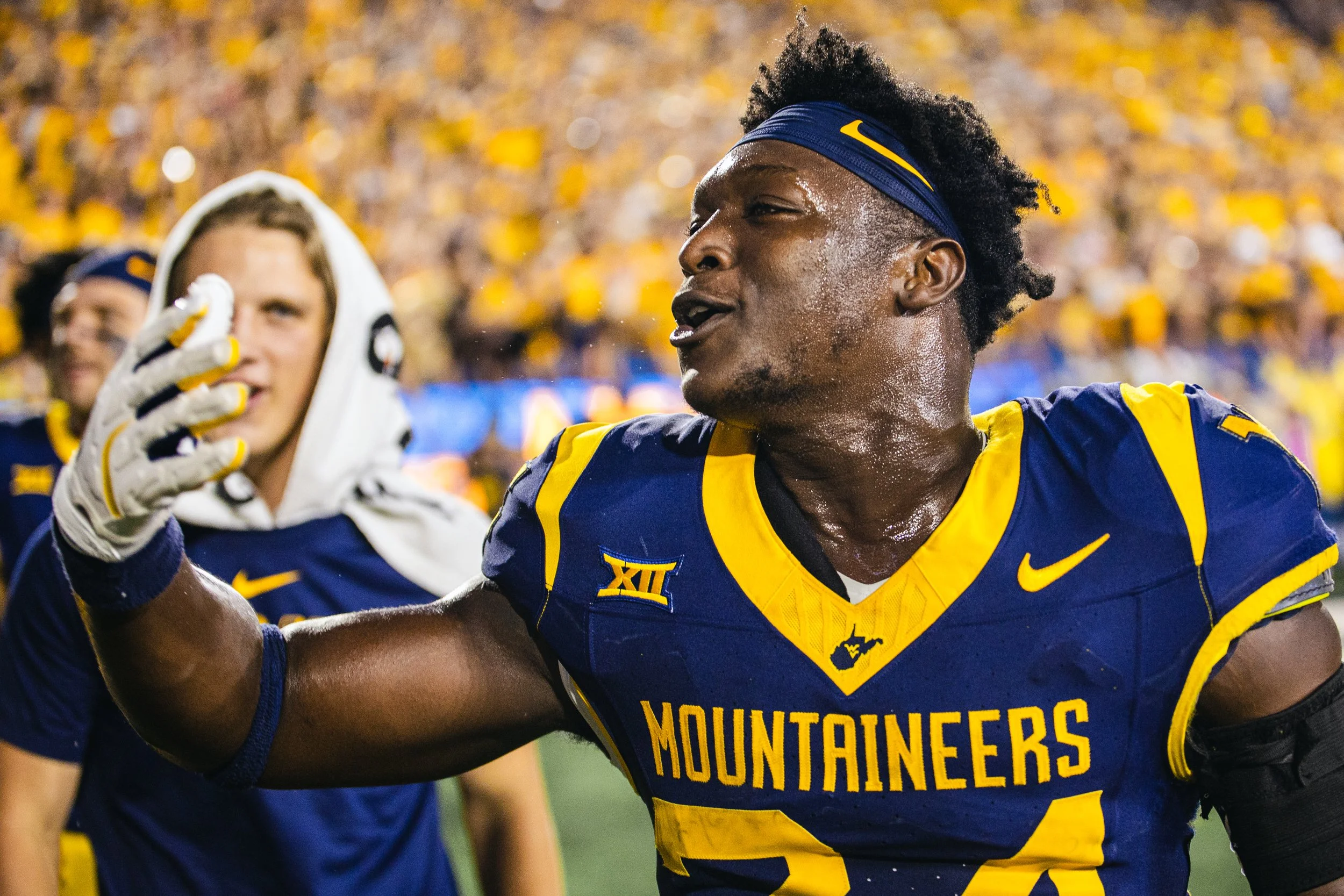 A football player in a blue and yellow uniform with WV state outline and 'MOUNTAINEERS' on the front, sweating, talking, and raising his arm. His teammate in the background has a white hoodie and gloves.