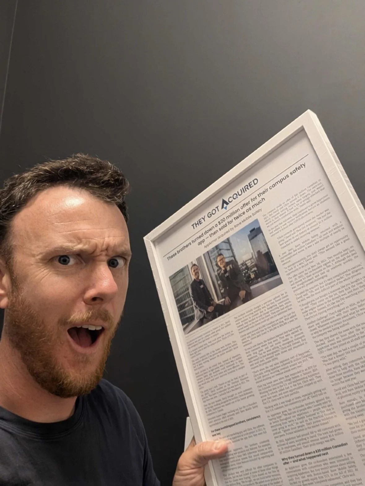 A man holding a framed article headlined They Got Acquired, in a white frame