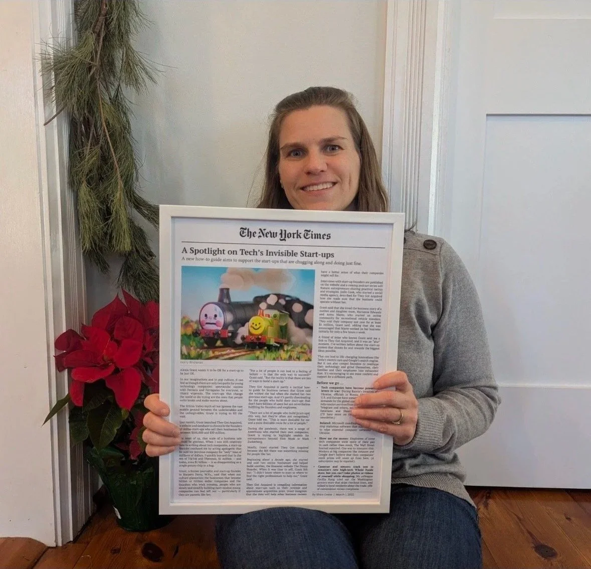 A woman smiling and holding her framed New York Times article about tech start-ups, in a white frame