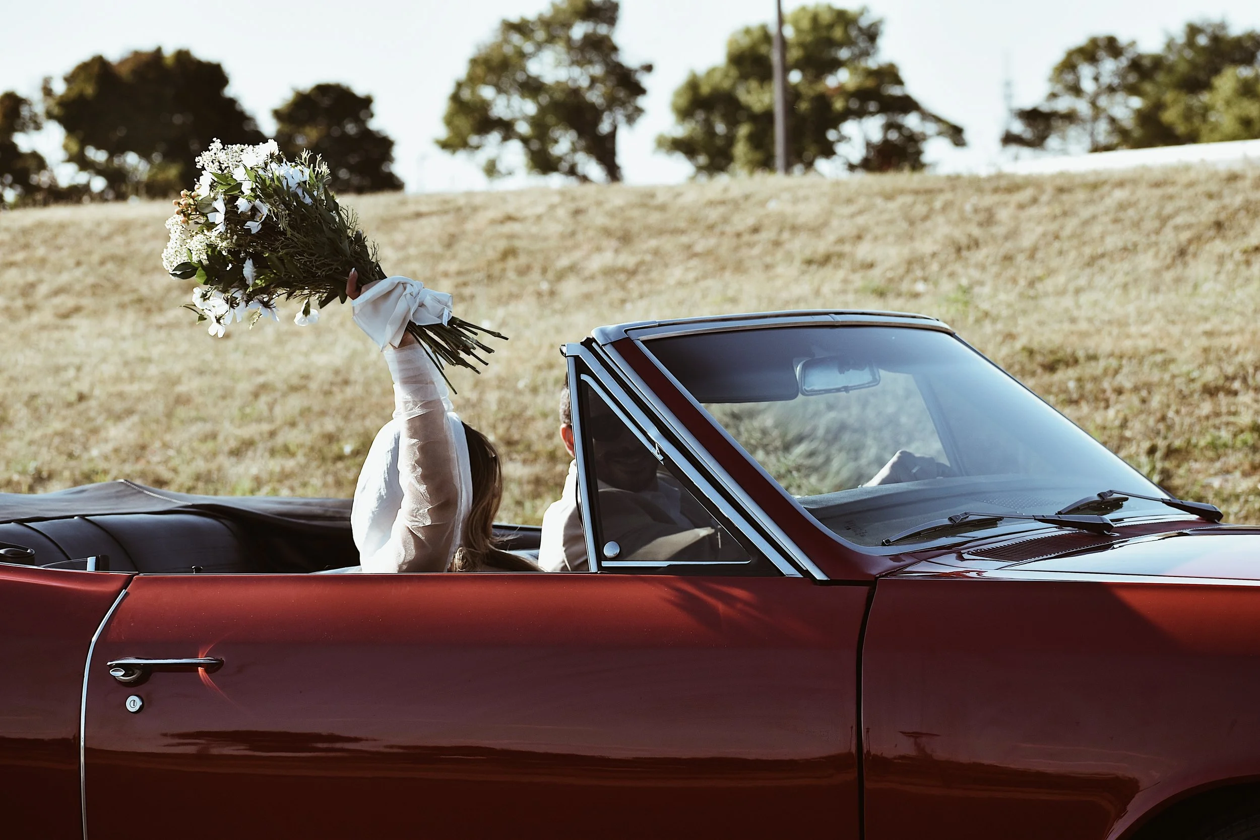 A woman in a white long-sleeve shirt holding a bouquet of flowers inside a red convertible car.