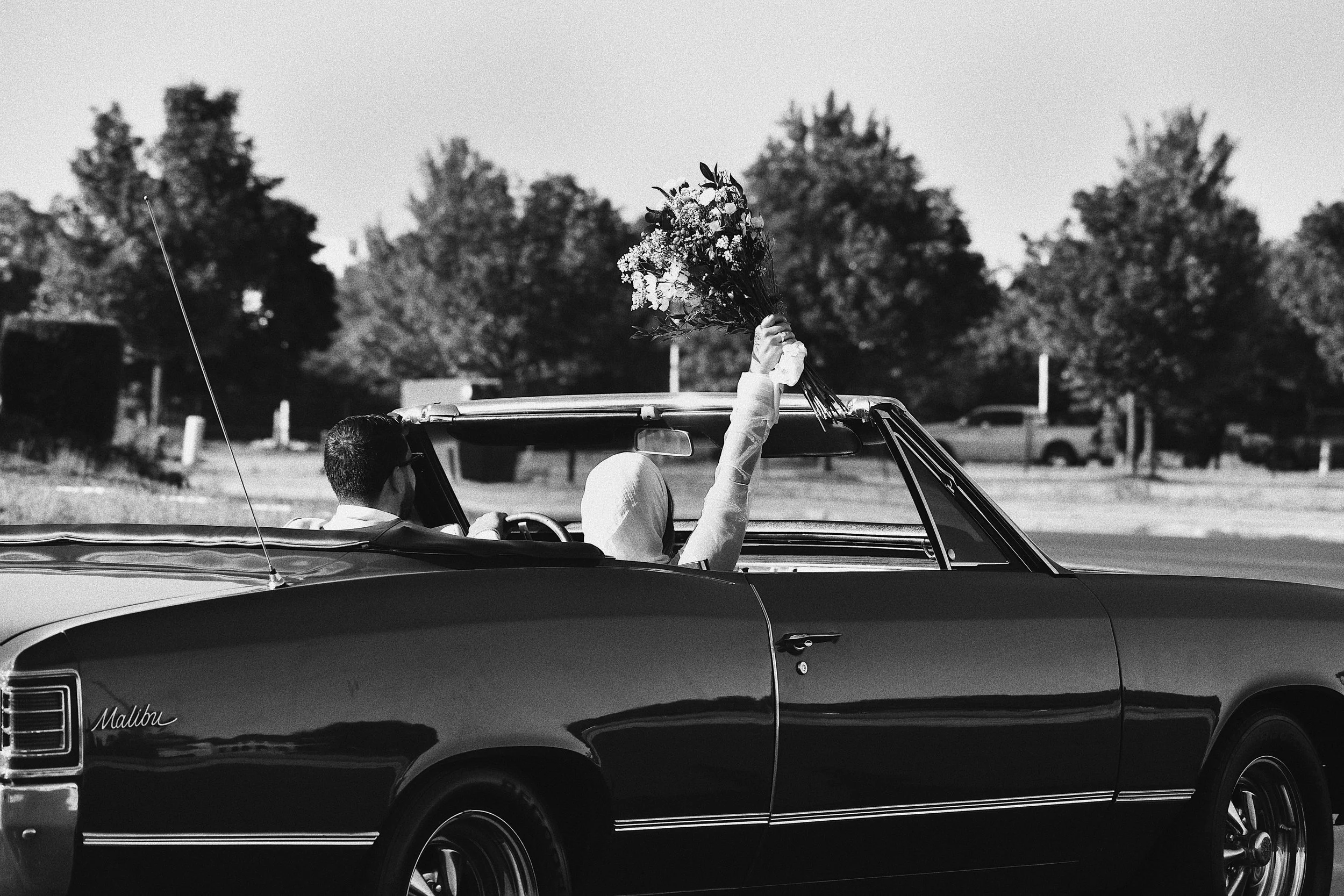 A black and white photo of two people in a vintage convertible car, one person holding a bouquet of flowers raised in the air.