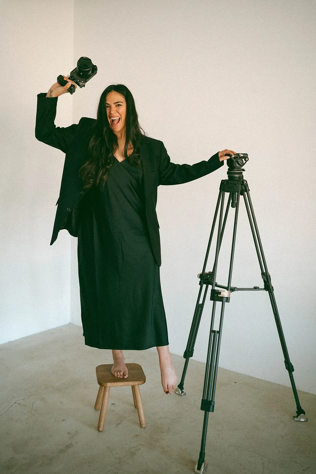 A woman in a black dress and blazer standing on a stool, holding a camera in her right hand and leaning on a tripod with a camera on it, joyfully posing in a minimal studio with plain walls.