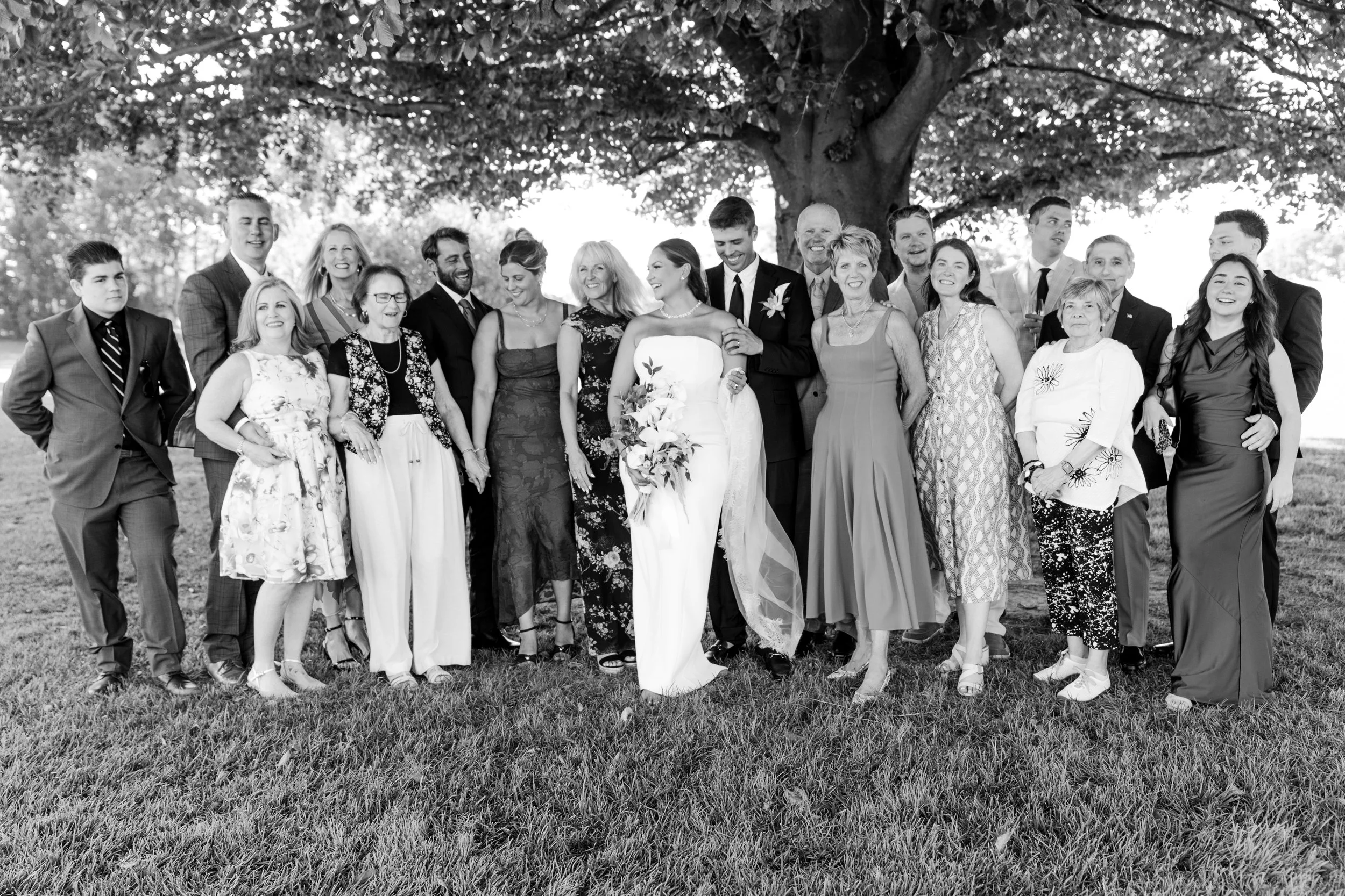 Black and white photo of a group of people at a wedding, standing outdoors in front of a large tree, dressed in formal attire, with the bride and groom in the center. documentary style wedding videographer based in new jersey