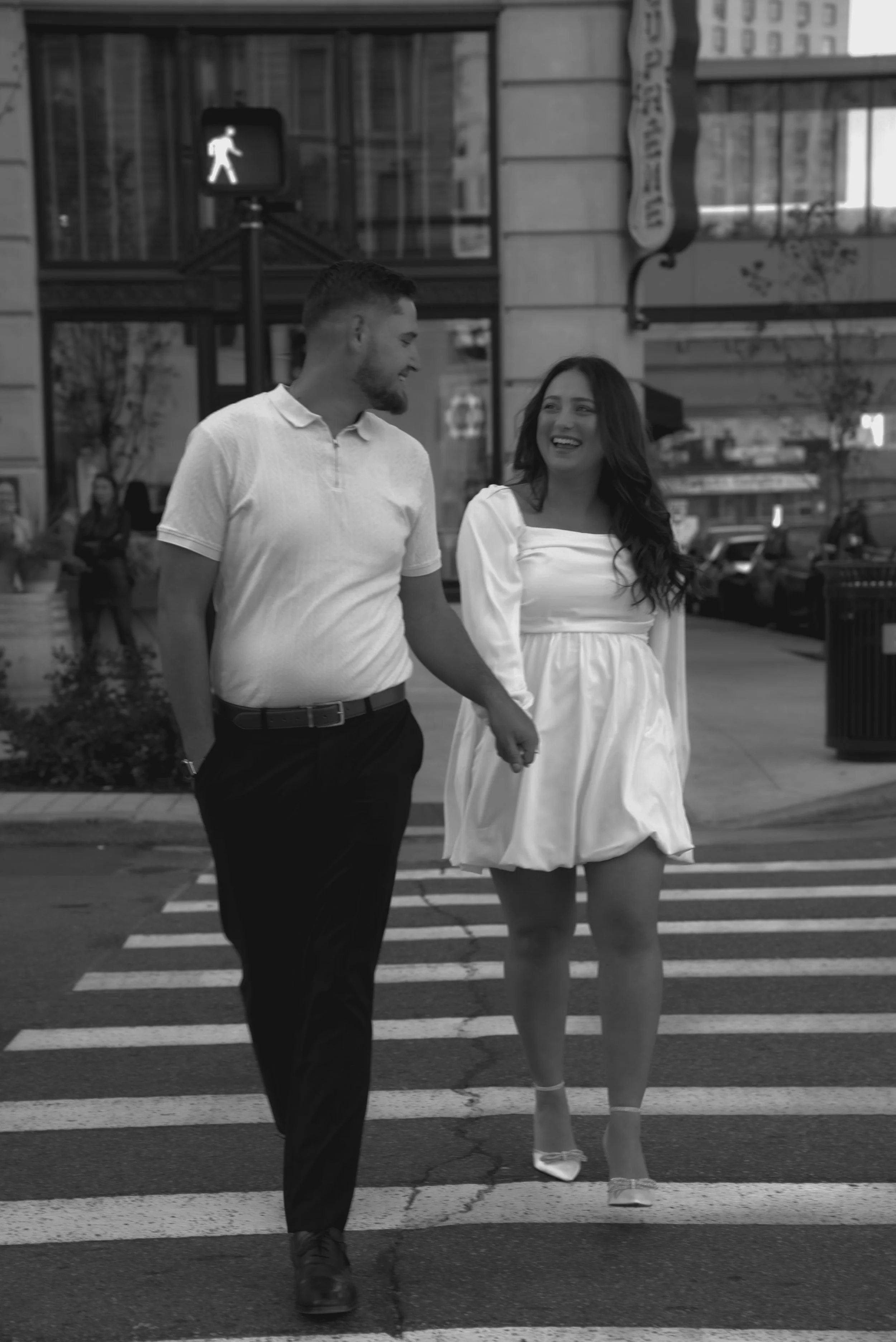 A man and woman walking hand in hand across a city crosswalk, smiling at each other, with traffic and buildings in the background.