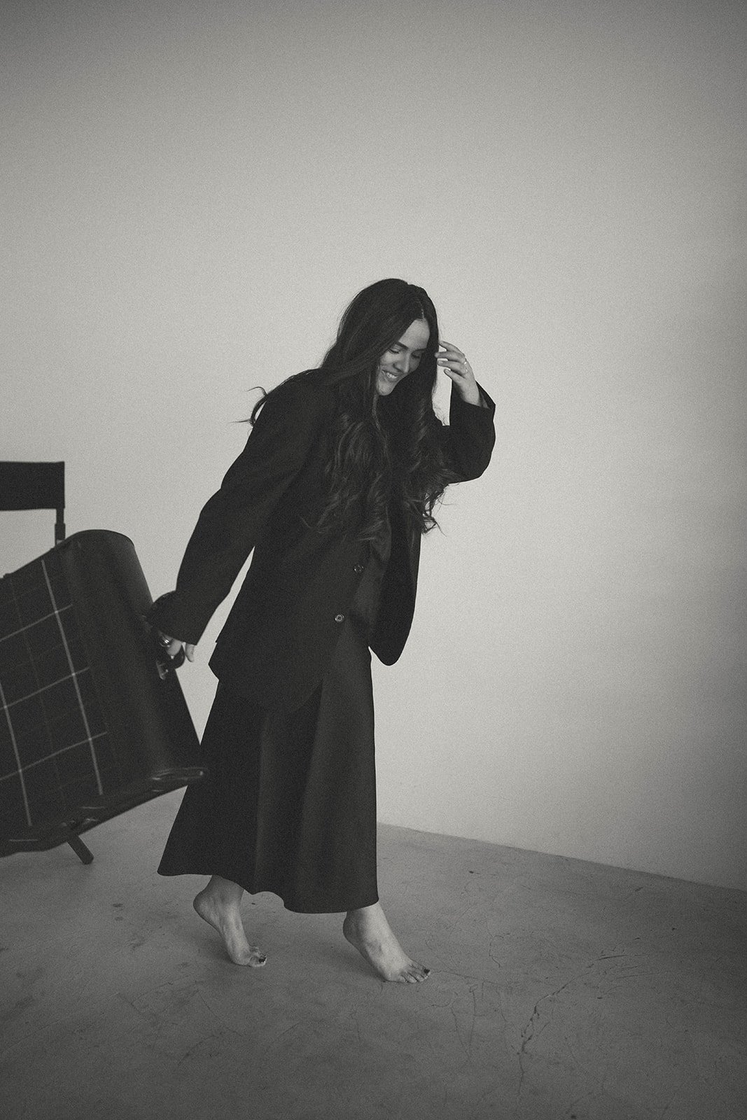 A woman with long dark hair, dressed in black, is smiling and barefoot, standing on a cracked floor, holding a plaid suitcase, against a plain white wall.