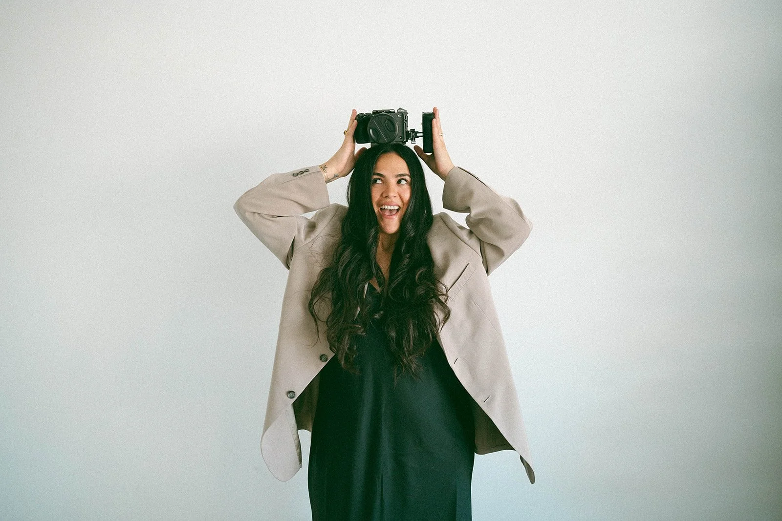 Woman with long dark hair wearing a beige blazer and black dress, holding a camera above her head, smiling in front of a plain light-colored wall.
