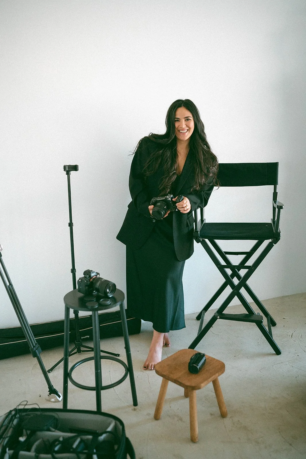 A woman with long dark hair in a black blazer and dress smiling while holding a camera in a photography studio with equipment around her.