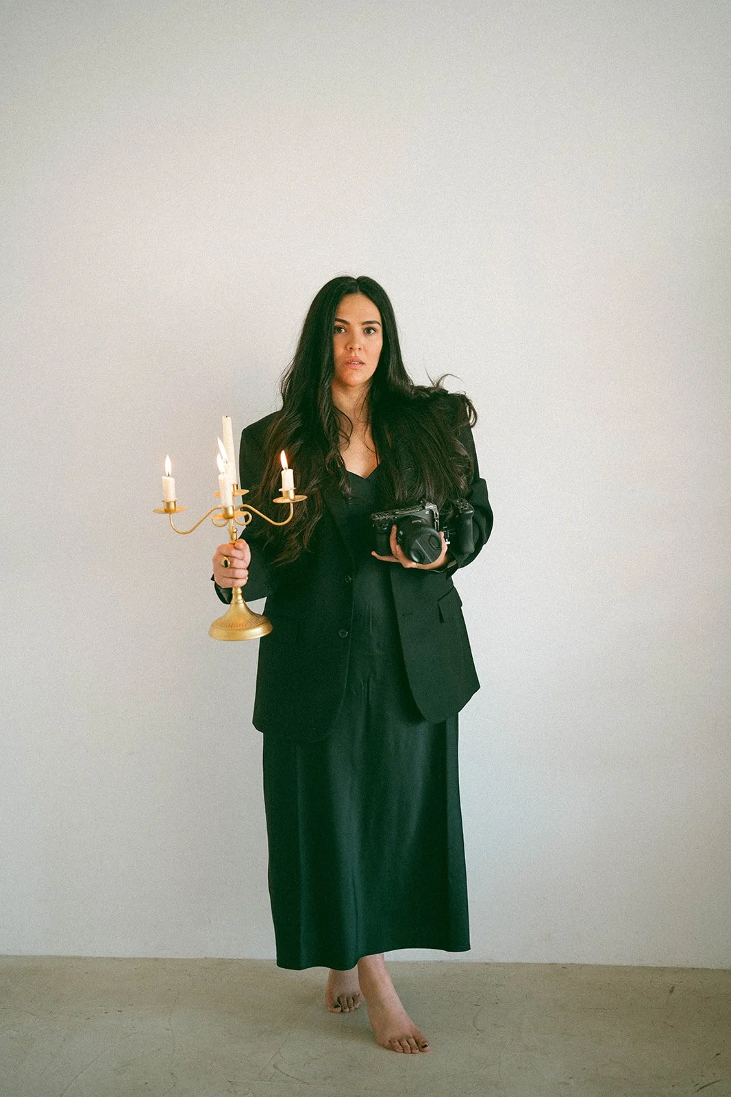 Woman with long dark hair, dressed in black blazer and skirt, standing barefoot against a plain white wall, holding a candlestick with three lit candles in her right hand and a camera in her left hand.