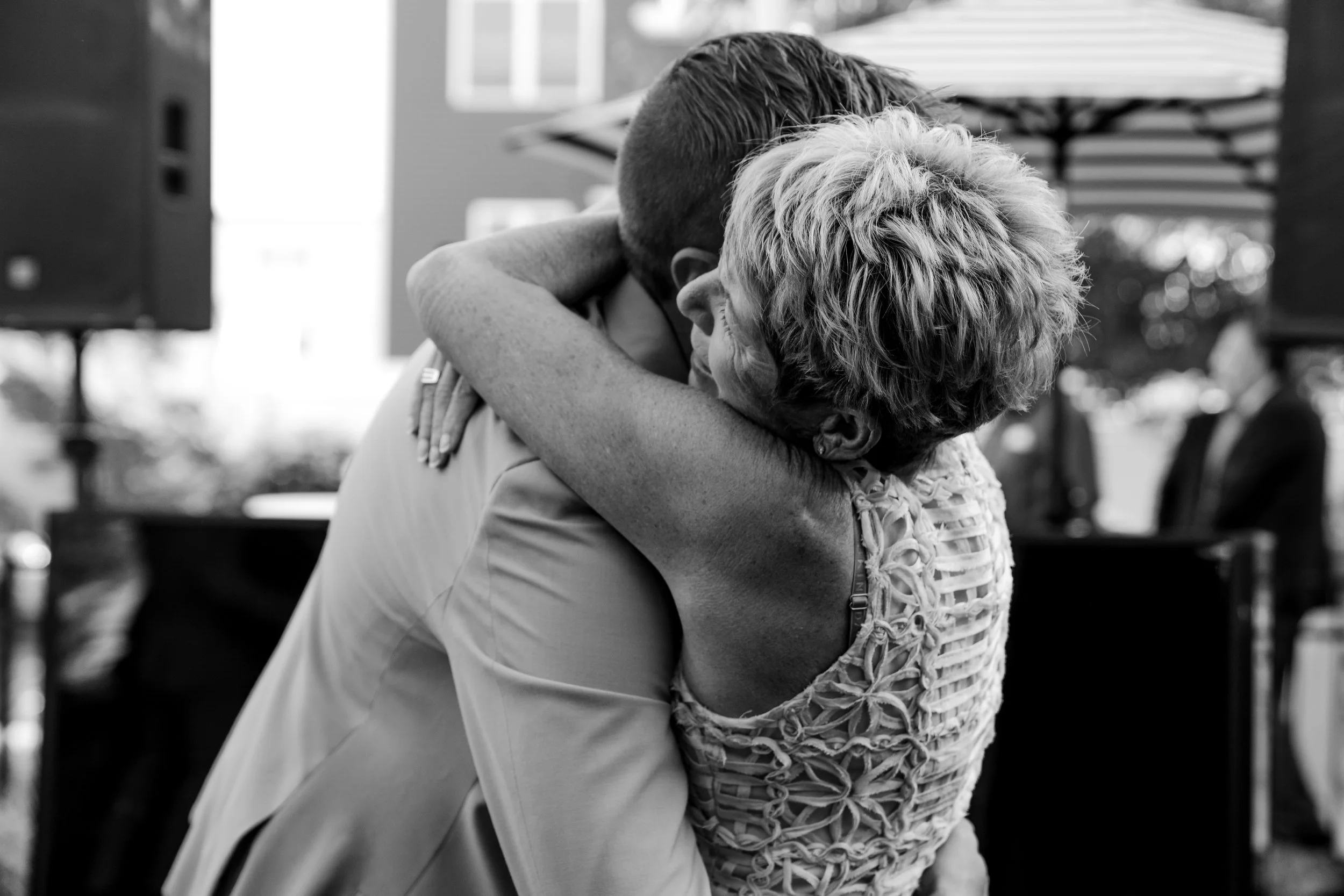 A black-and-white photo of a man and woman hugging tightly, with only part of their faces and arms visible. The woman has short, curly hair and is wearing a lace or crochet top. They are outdoors, with blurred background including a parasol and trees.