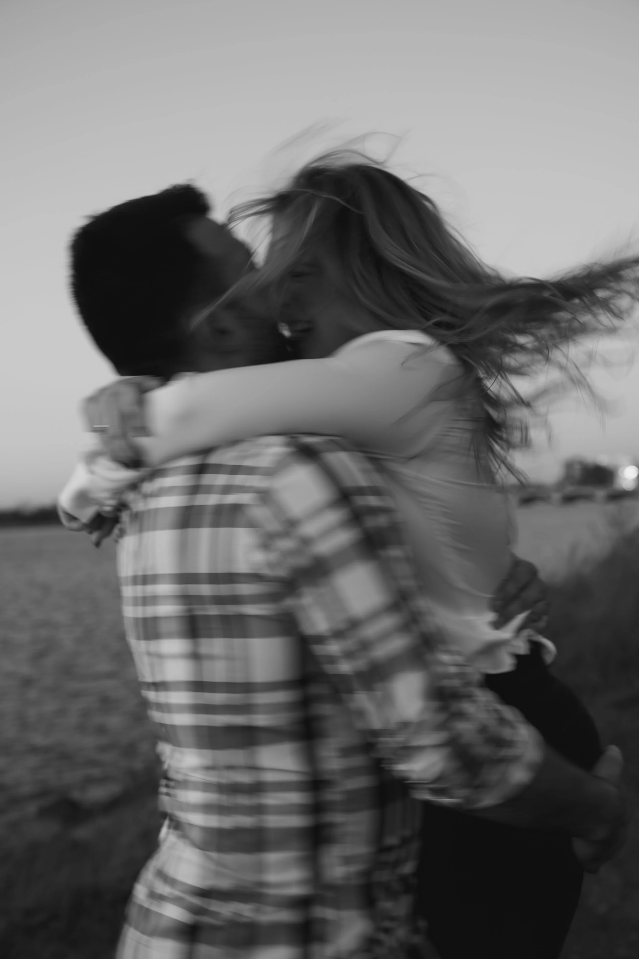 A couple sharing an embrace outdoors, with the woman smiling and her hair flowing, captured in black and white.