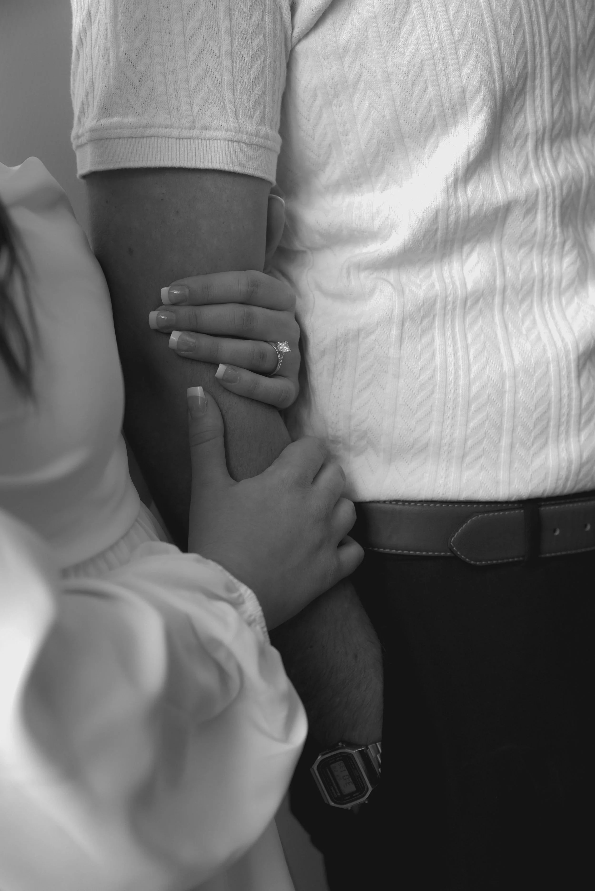 A close-up black-and-white photo of a person holding a child's arm, with the child's hand resting on the person's wrist. The person has a wedding ring on their finger and is wearing a patterned shirt and a belt with a watch on their wrist.