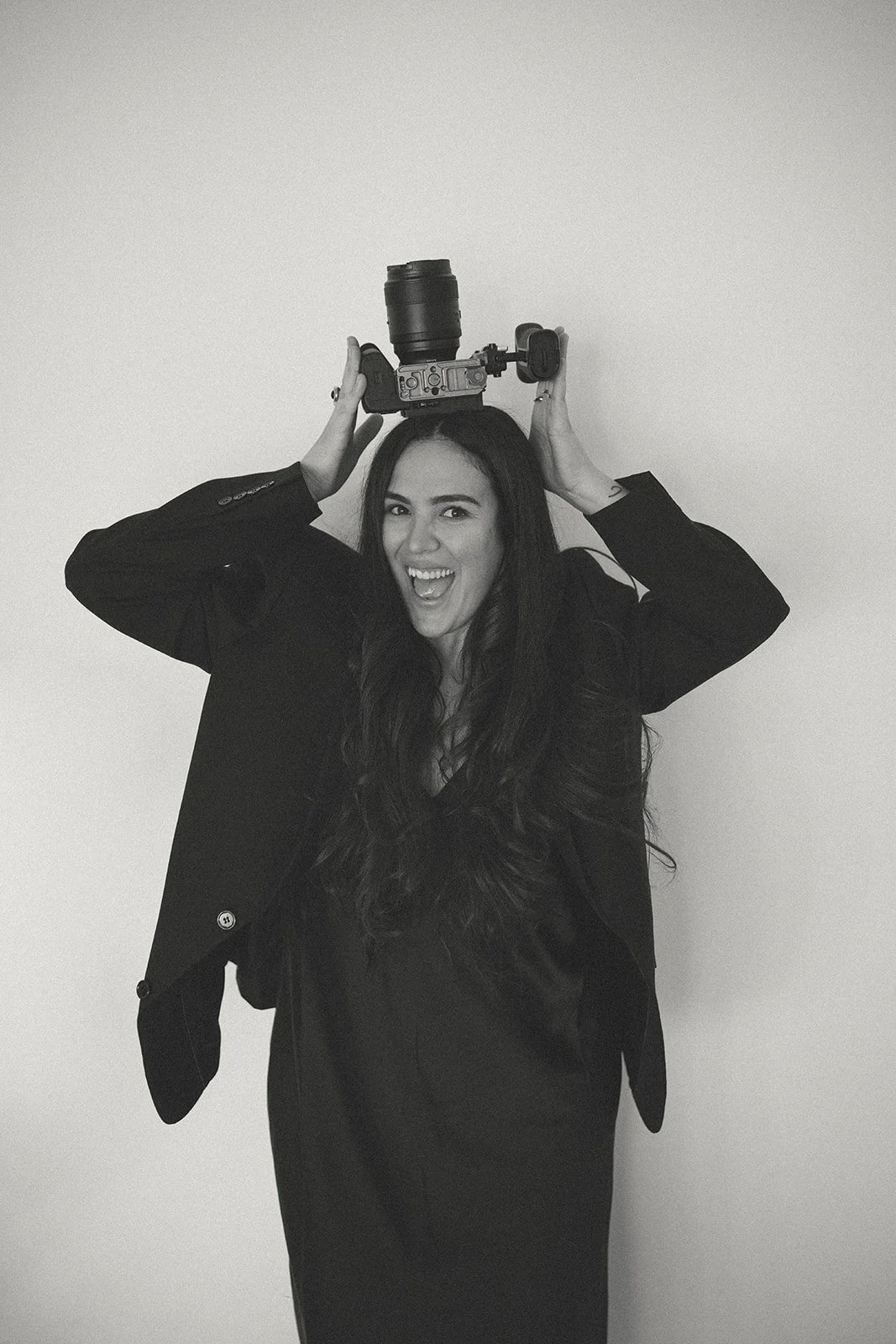 Lettywayfilms New Jersey wedding videographer with long dark hair holding a camera on her head, smiling and winking, wearing a black blazer and dress, standing against a plain wall.