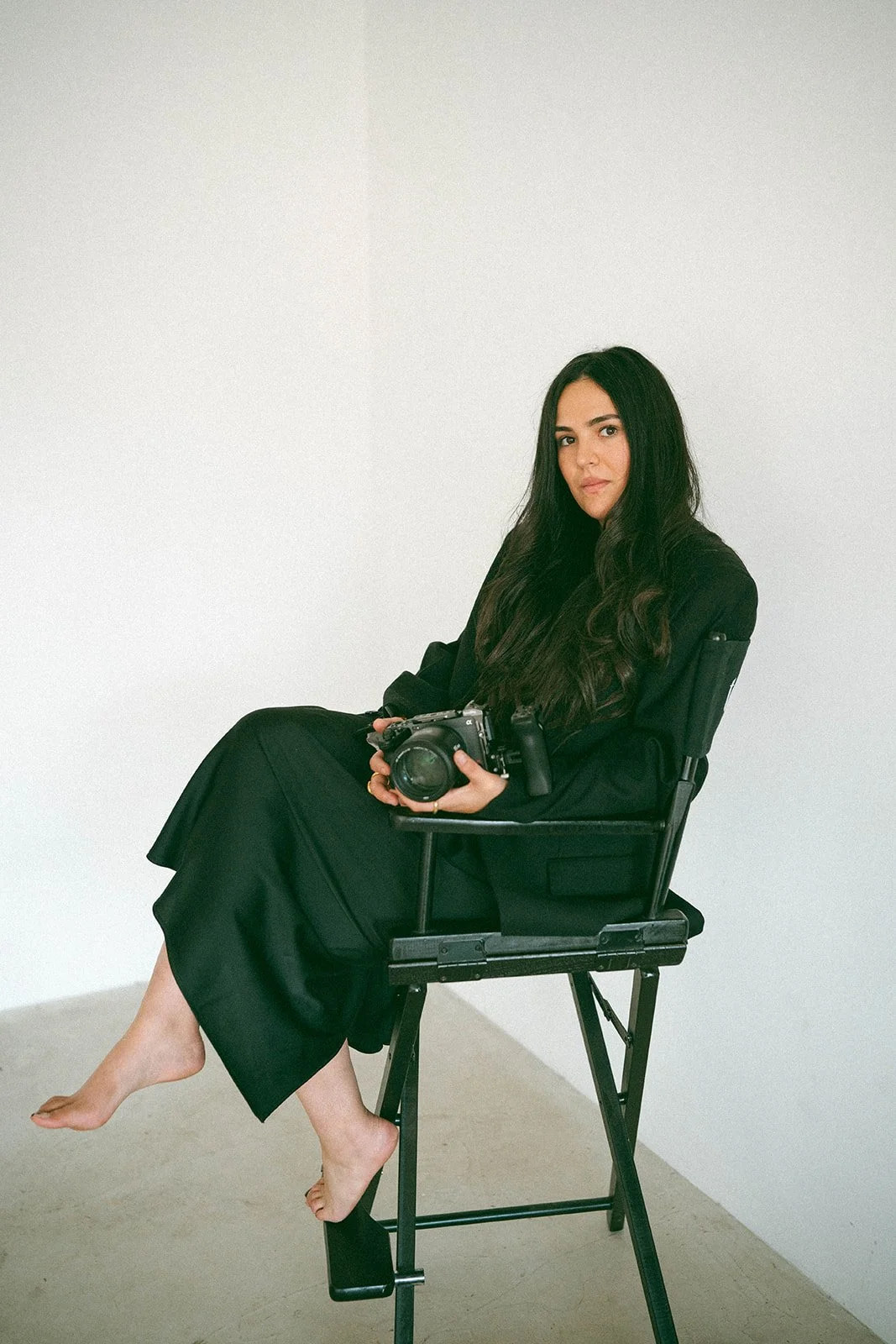 A woman with long dark hair sitting barefoot on a chair with a camera in her hands against a plain white wall.