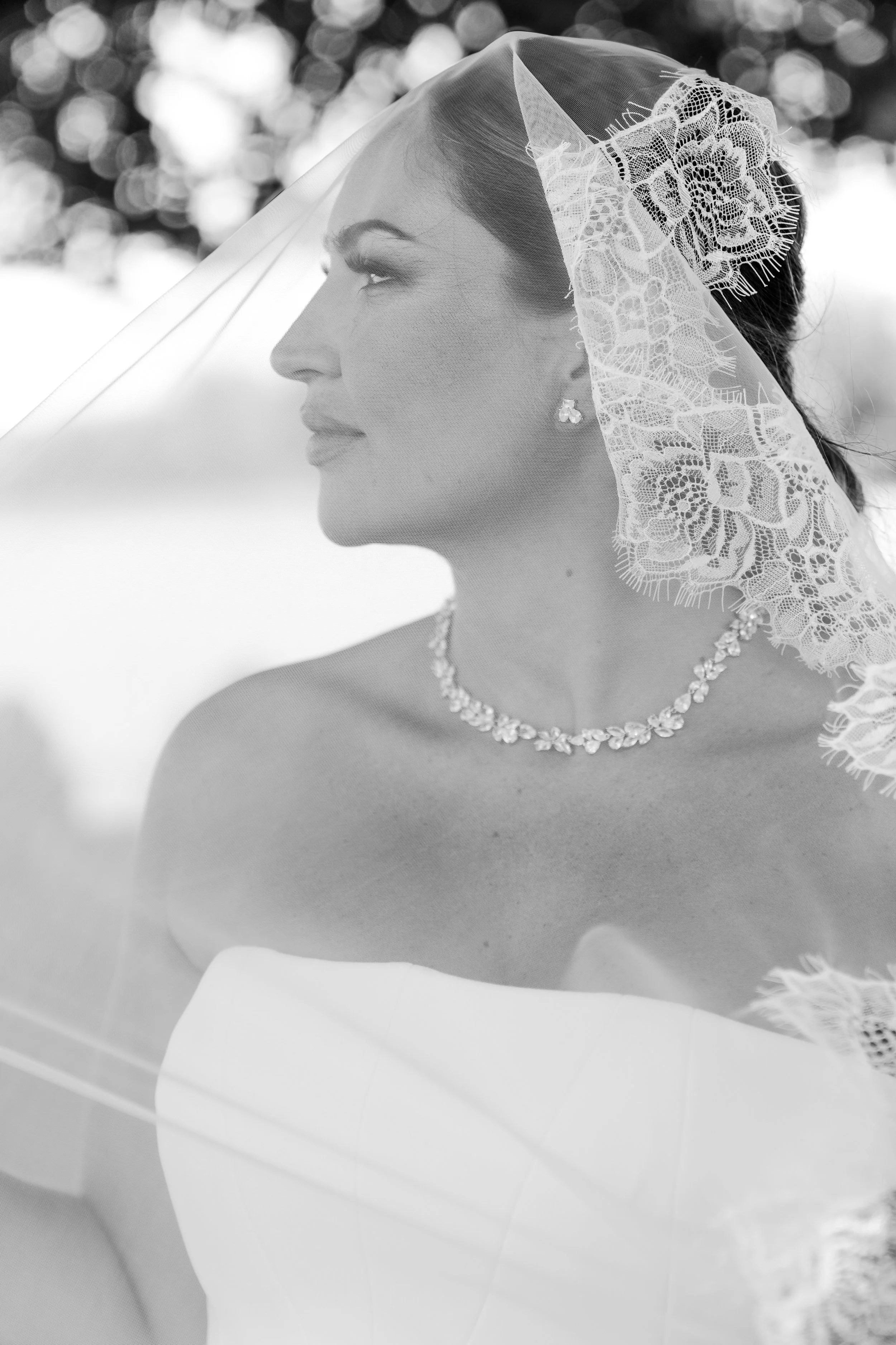 Black and white photo of a bride in a wedding dress with a lace veil, jewelry, and earrings, looking to the left.