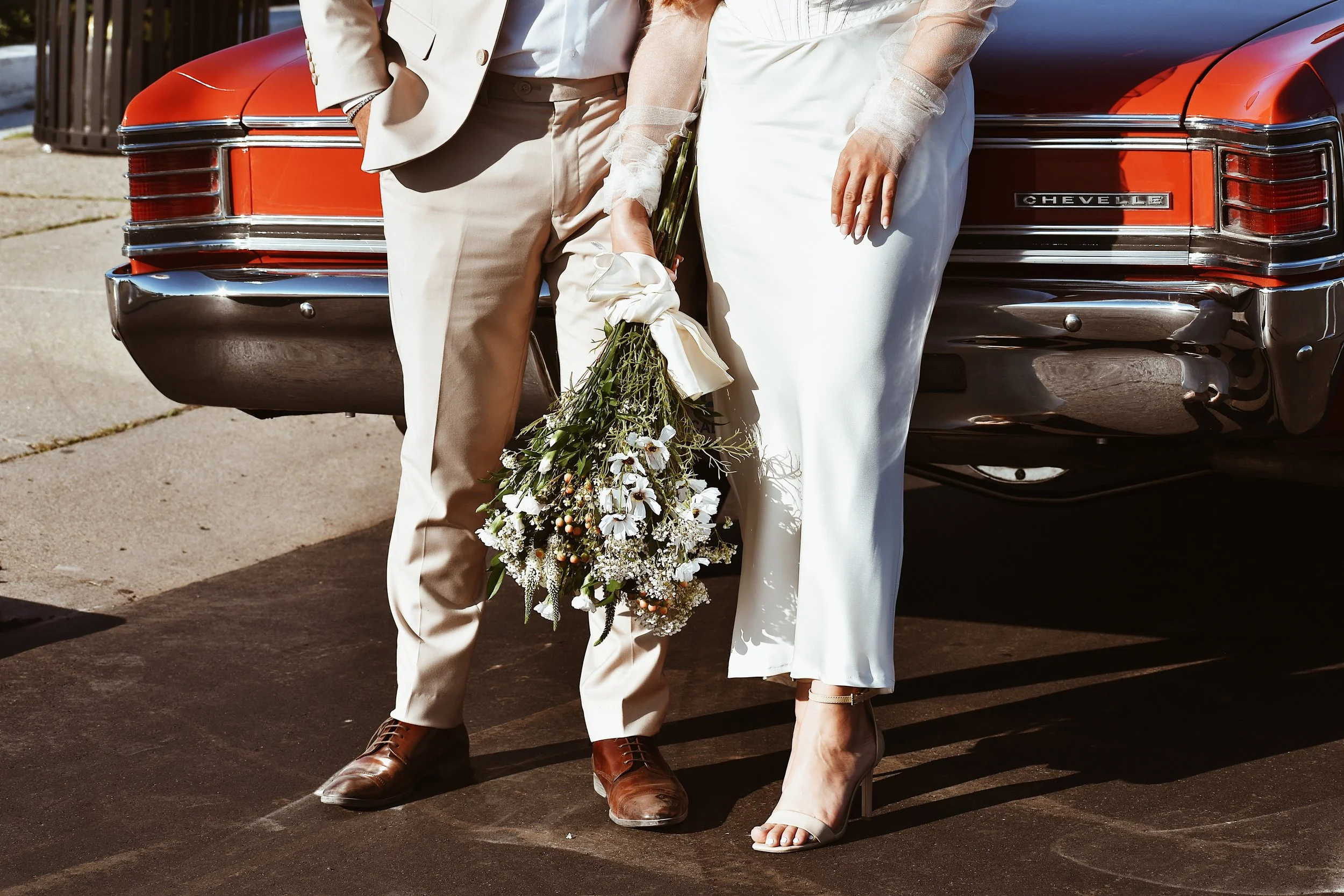 Close-up of a bride and groom standing in front of a red Chevrolet car on their wedding day, with the bride holding a bouquet of white and green flowers.