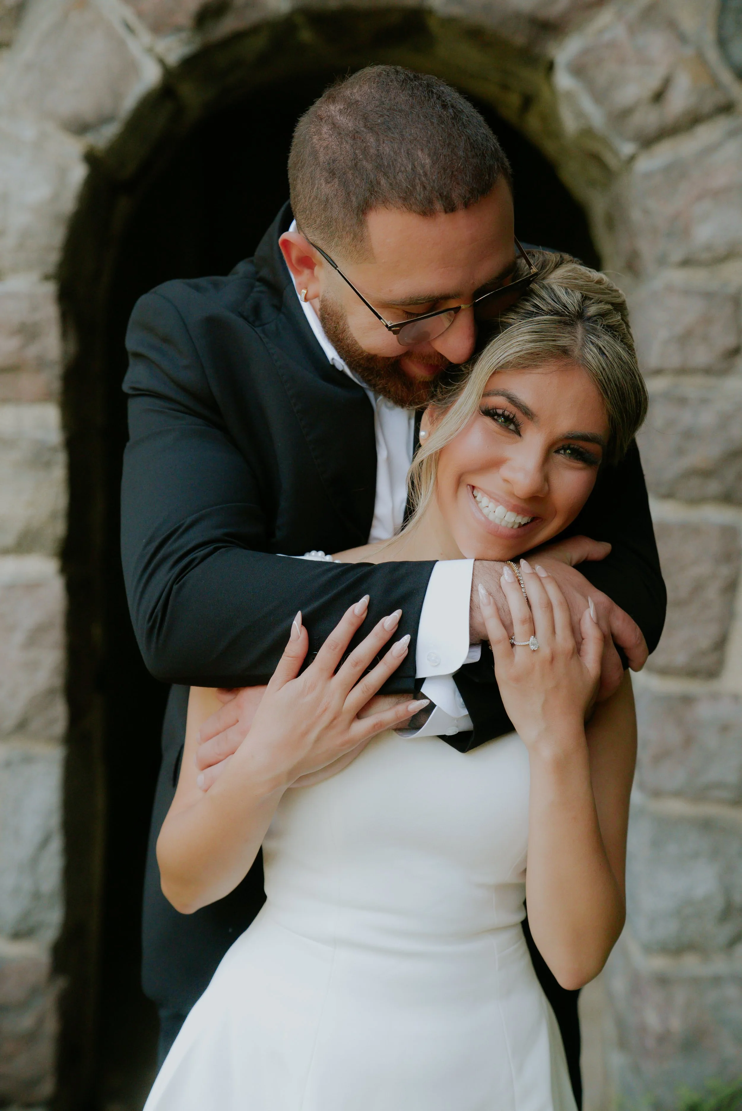 A smiling bride and groom embrace outdoors, with the groom wearing glasses and a black suit and the bride in a white dress showing her ring. Inspired by wedding videography by lettywayfilms