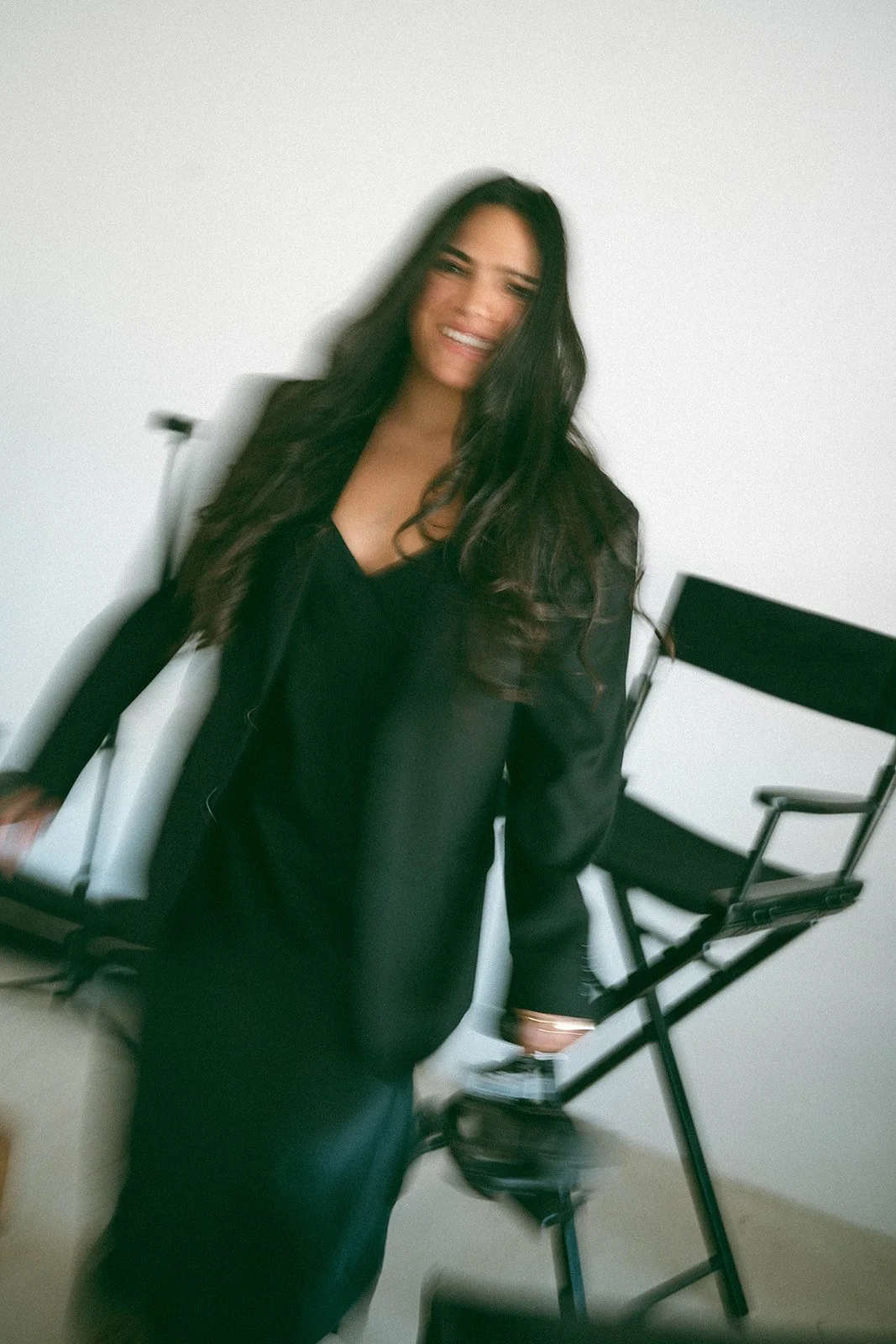 A woman with long dark hair smiling, standing in front of white background, wearing black clothing.