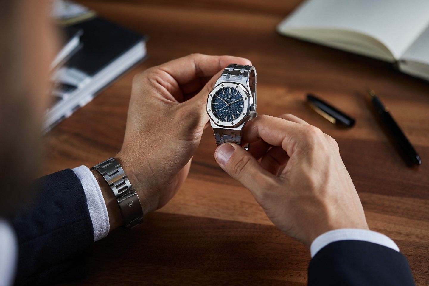 Person holding luxury wristwatch with blue dial and silver bracelet, sitting at a wooden desk with notebooks and a pen.