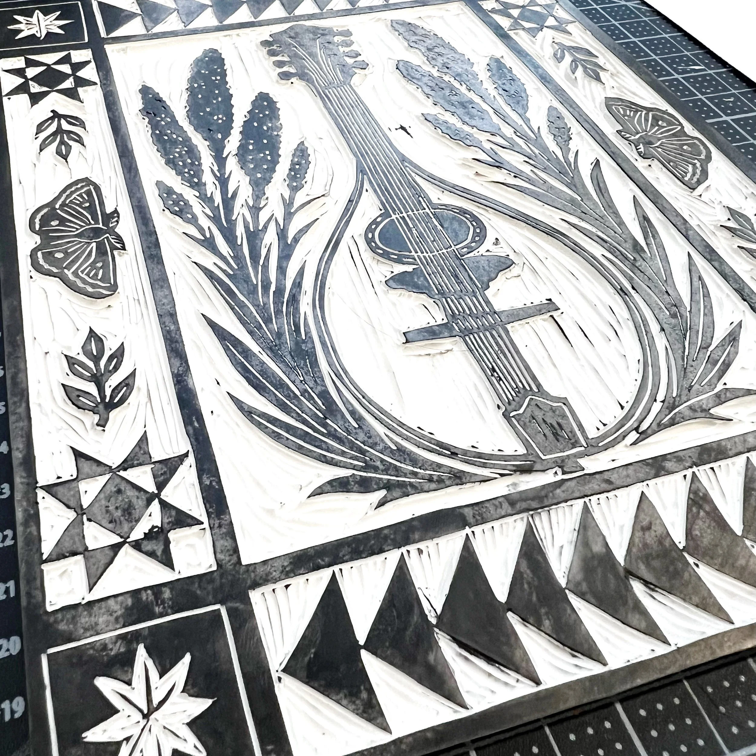 Work-in-progress linocut block featuring an octavve mandolin surrounded by botanical motifs and quilt-inspired borders.