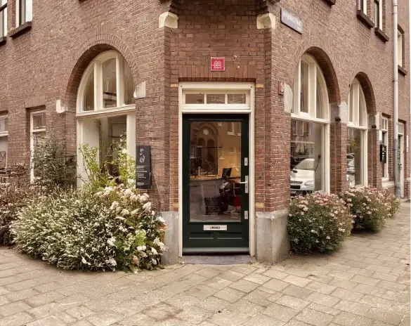 Een straathoek met een rode bakstenen gebouw, een groene deur, en drie grote ramen met witte kozijnen. Bloemen en planten groeien langs de gevel.