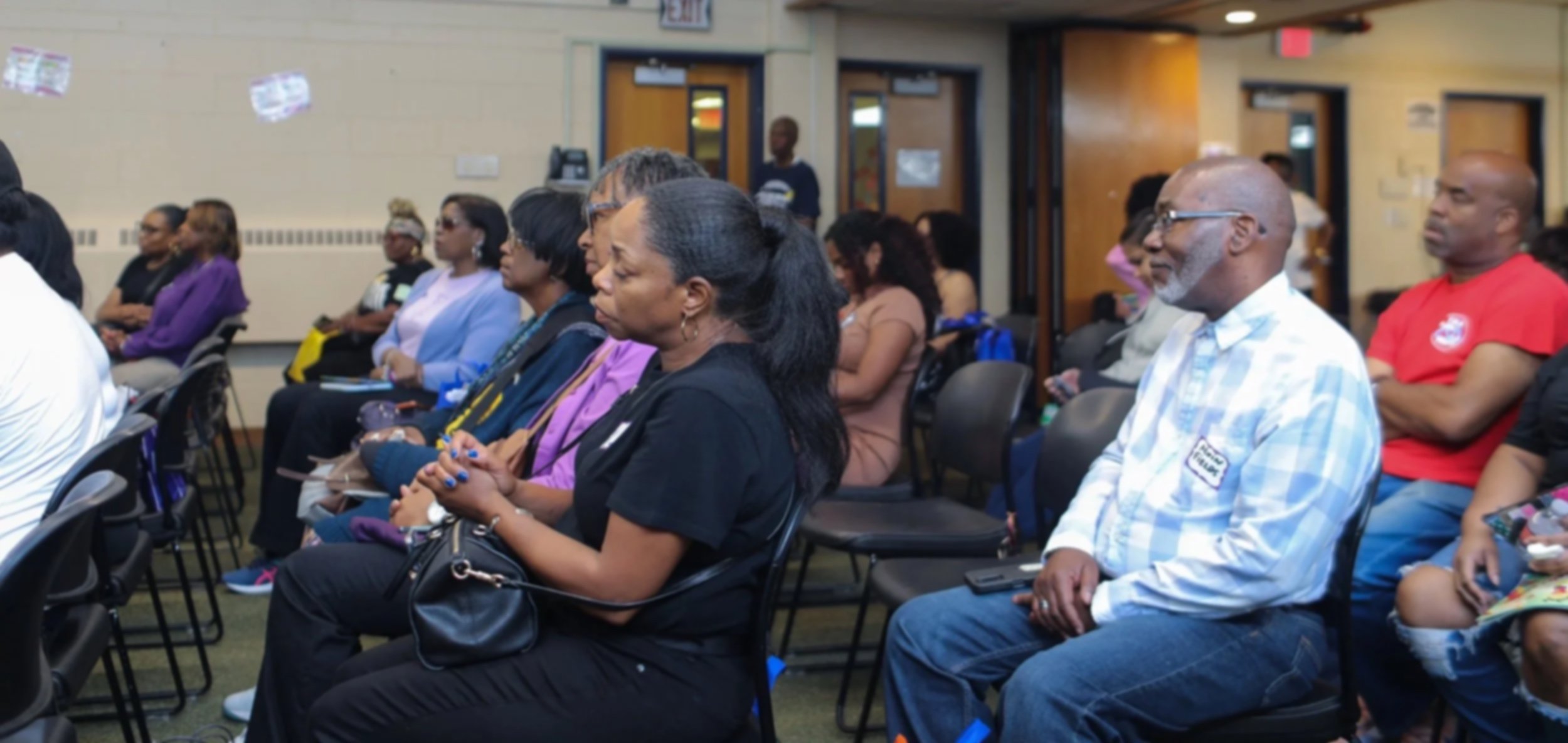 A group of diverse people seated in chairs attending a presentation or event in a room with beige walls and wooden doors.