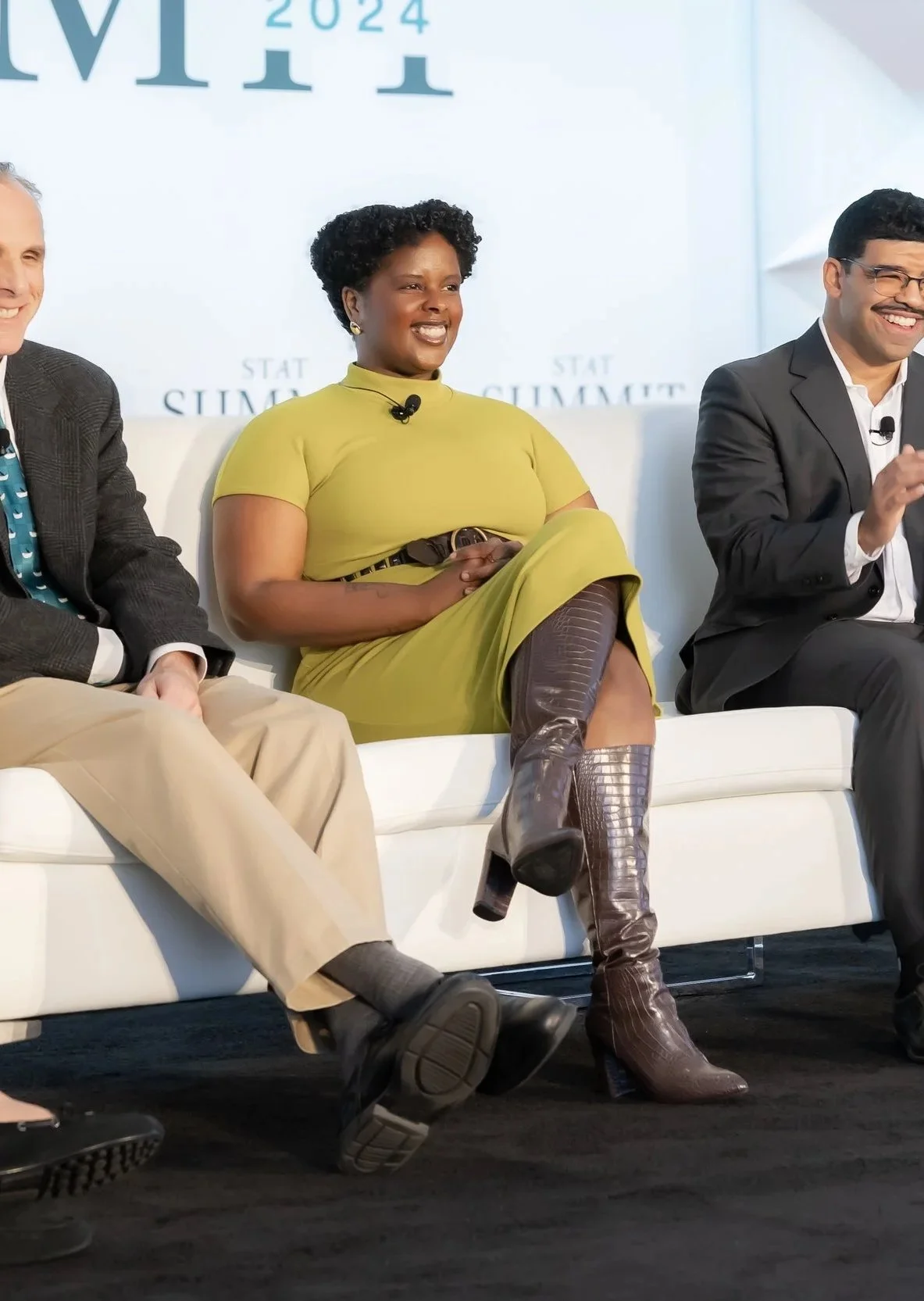Three people sitting on a white couch at a conference with a light blue background that says '2024' and 'STAT SUMMIT' behind them. The woman in the center is wearing a yellow dress and brown boots, smiling and looking to her right. The man on the right is wearing a black suit and glasses, also smiling. The person on the left is partially visible.