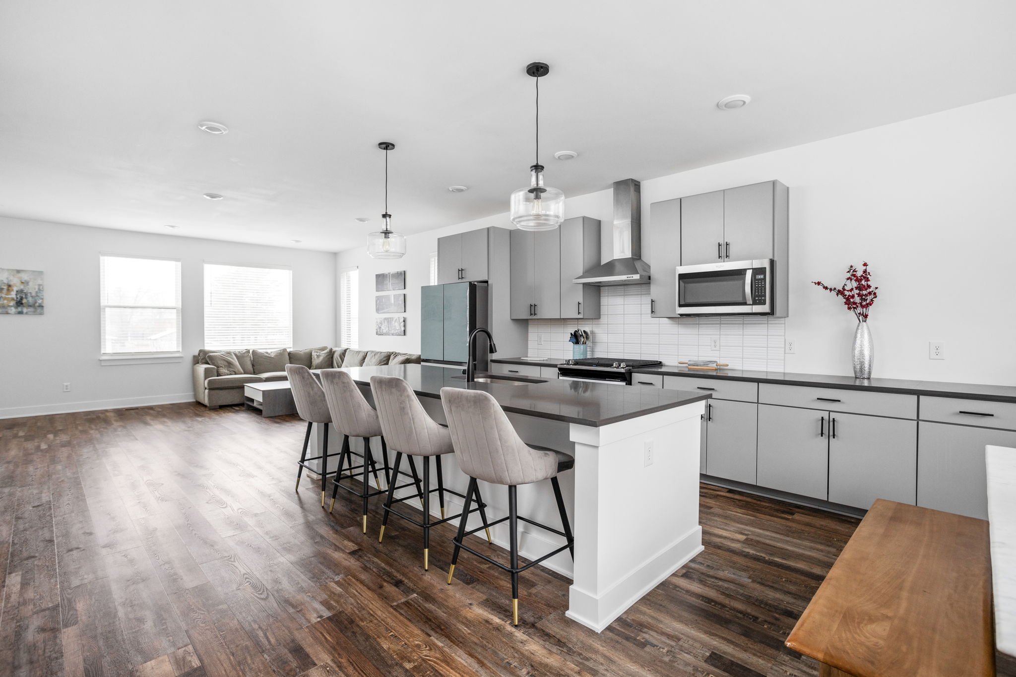 Modern kitchen with gray cabinets, island with stools, stainless steel appliances, and wood flooring. Open layout with living area and large windows.
