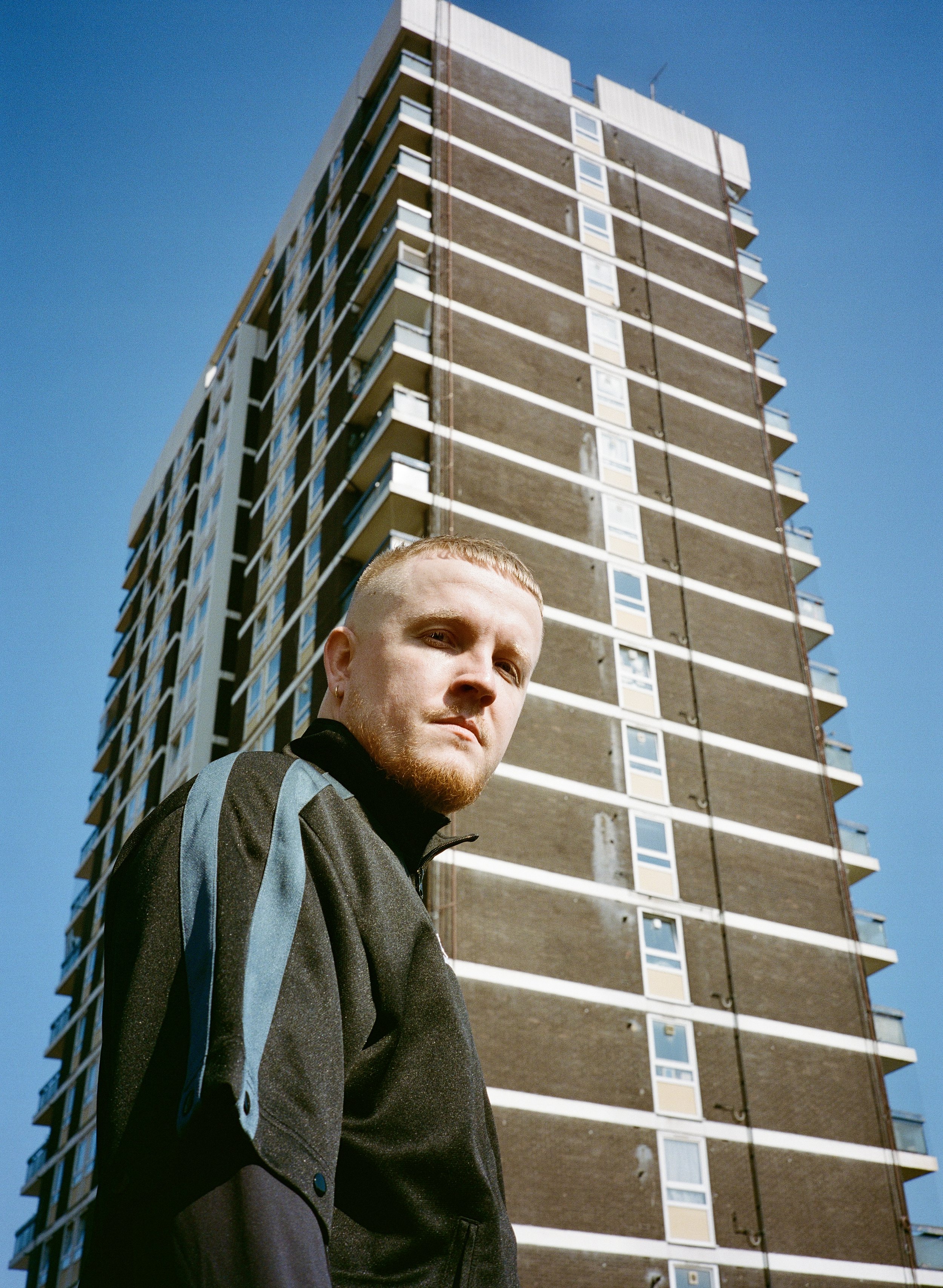 A young man with blond hair and a beard wearing a black and gray jacket, standing in front of a tall apartment building under a clear blue sky.