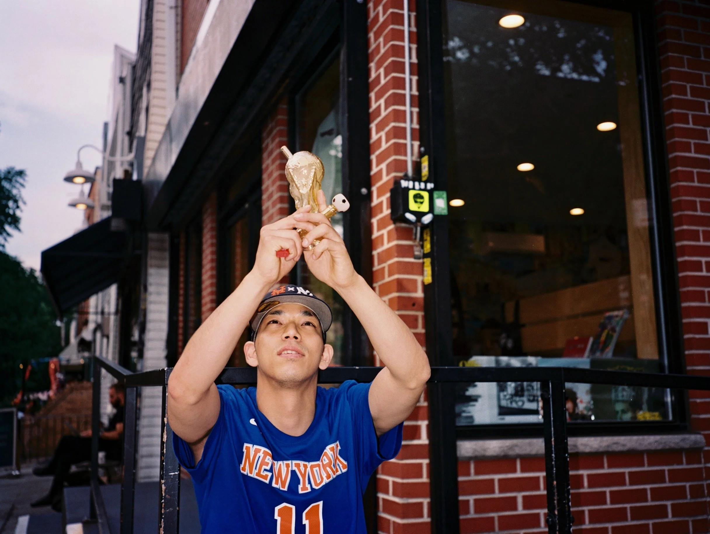 A person wearing a blue shirt with 'NEWYORK' written on it in orange and white letters and holding a plastic cup of ice cream with a spoon, standing behind a metal railing in front of a brick building with large windows at dusk.