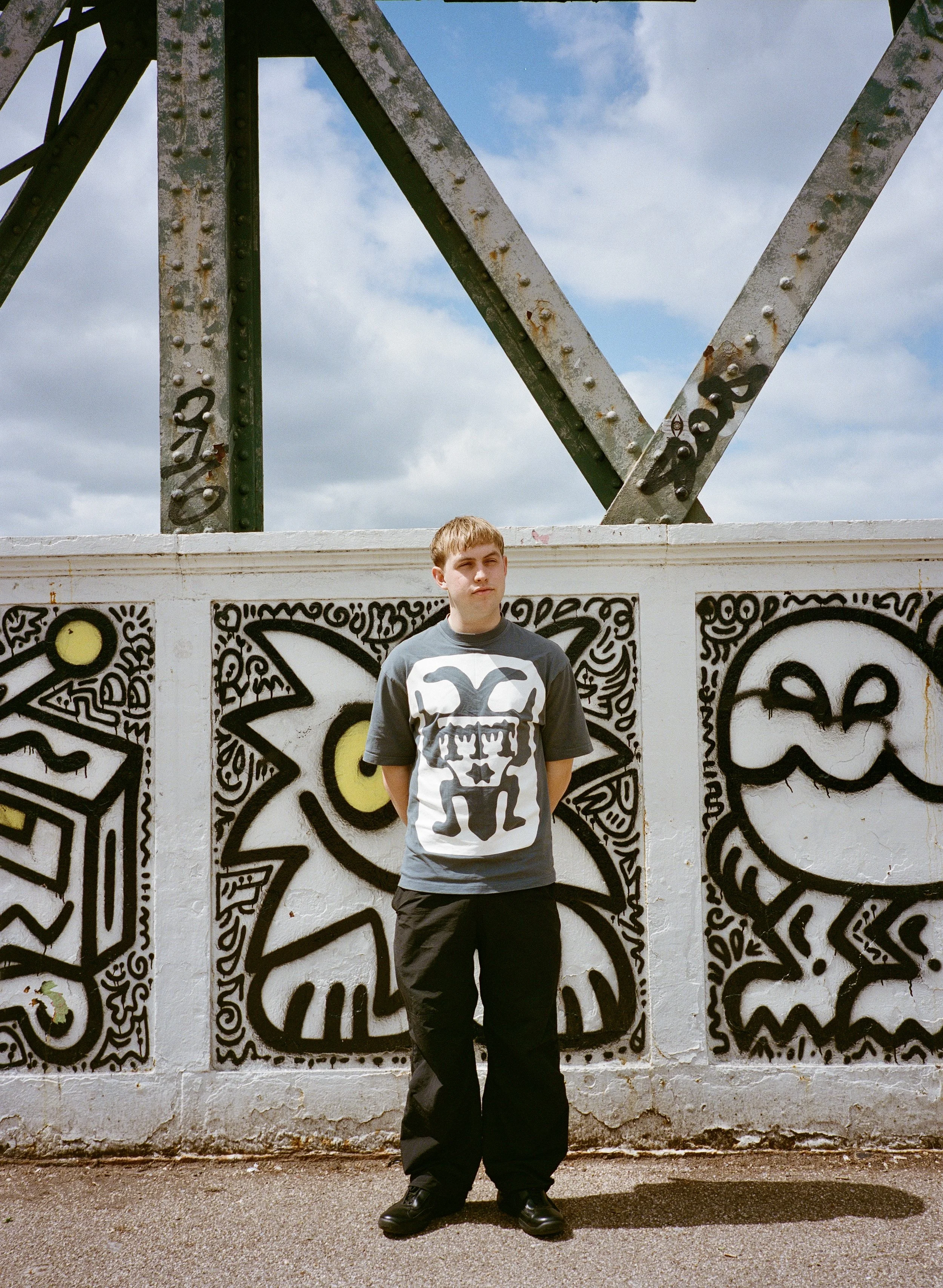Young man standing in front of a graffiti-covered wall and a metal structure with a partly cloudy sky in the background.