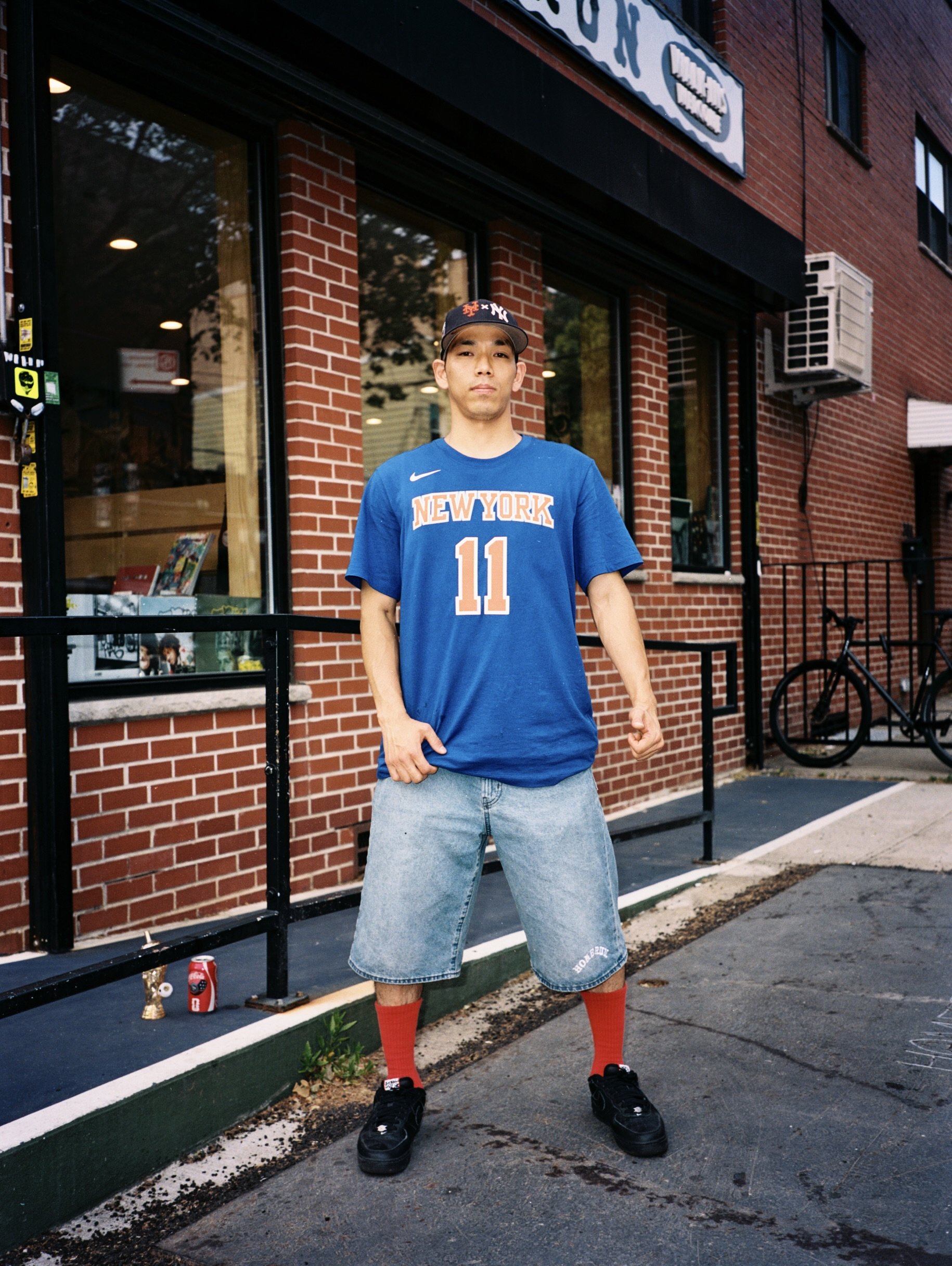 A young man in a blue New York basketball jersey number 11, wearing gray shorts, red socks, and black shoes, standing outside a brick building with large windows and a sign. There are personal items and a soda can on the ground nearby.