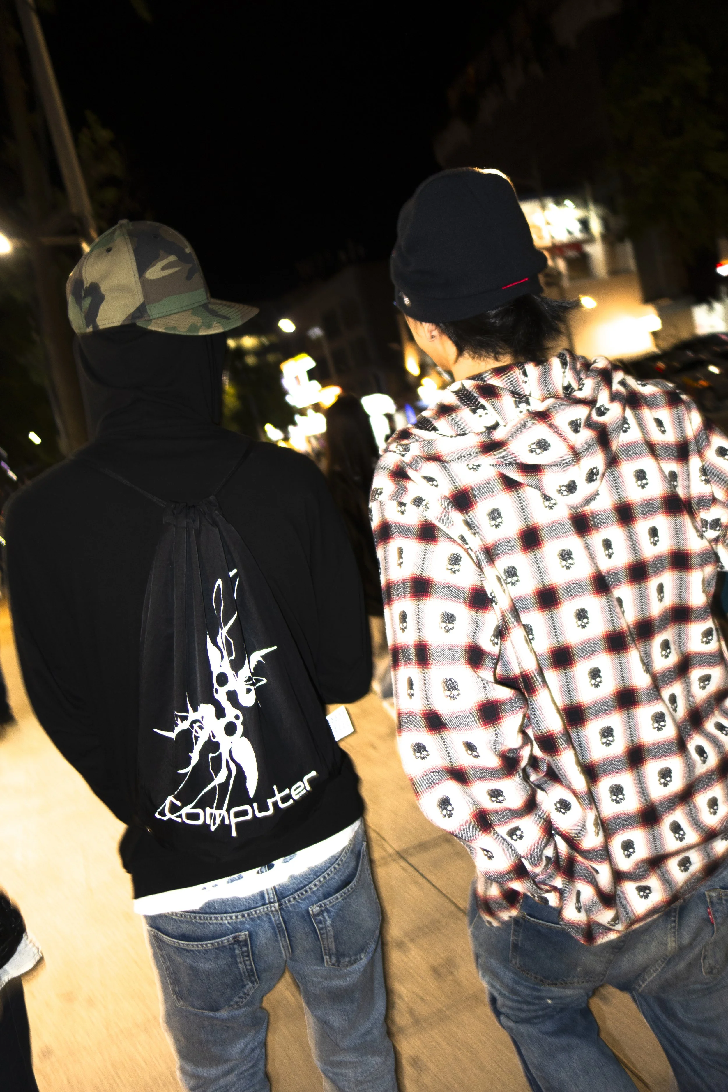 Two people standing side by side at night, seen from behind. The person on the left wears a black hoodie with white graphic design and a camouflage cap. The person on the right wears a black beanie and a plaid shirt with skull patterns.