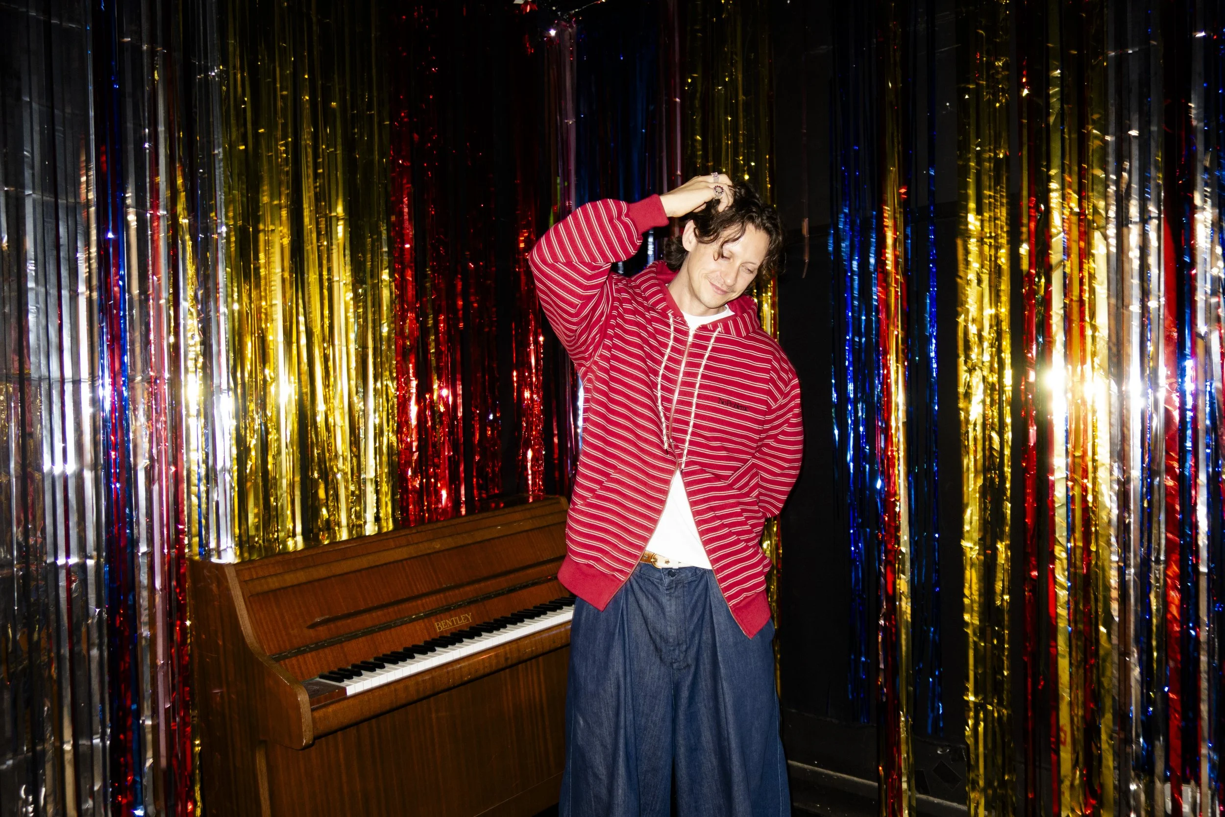 Person with curly hair dancing with eyes closed, wearing a red and white striped hoodie and blue jeans, standing in front of a colorful metallic fringe curtain and a wooden piano.
