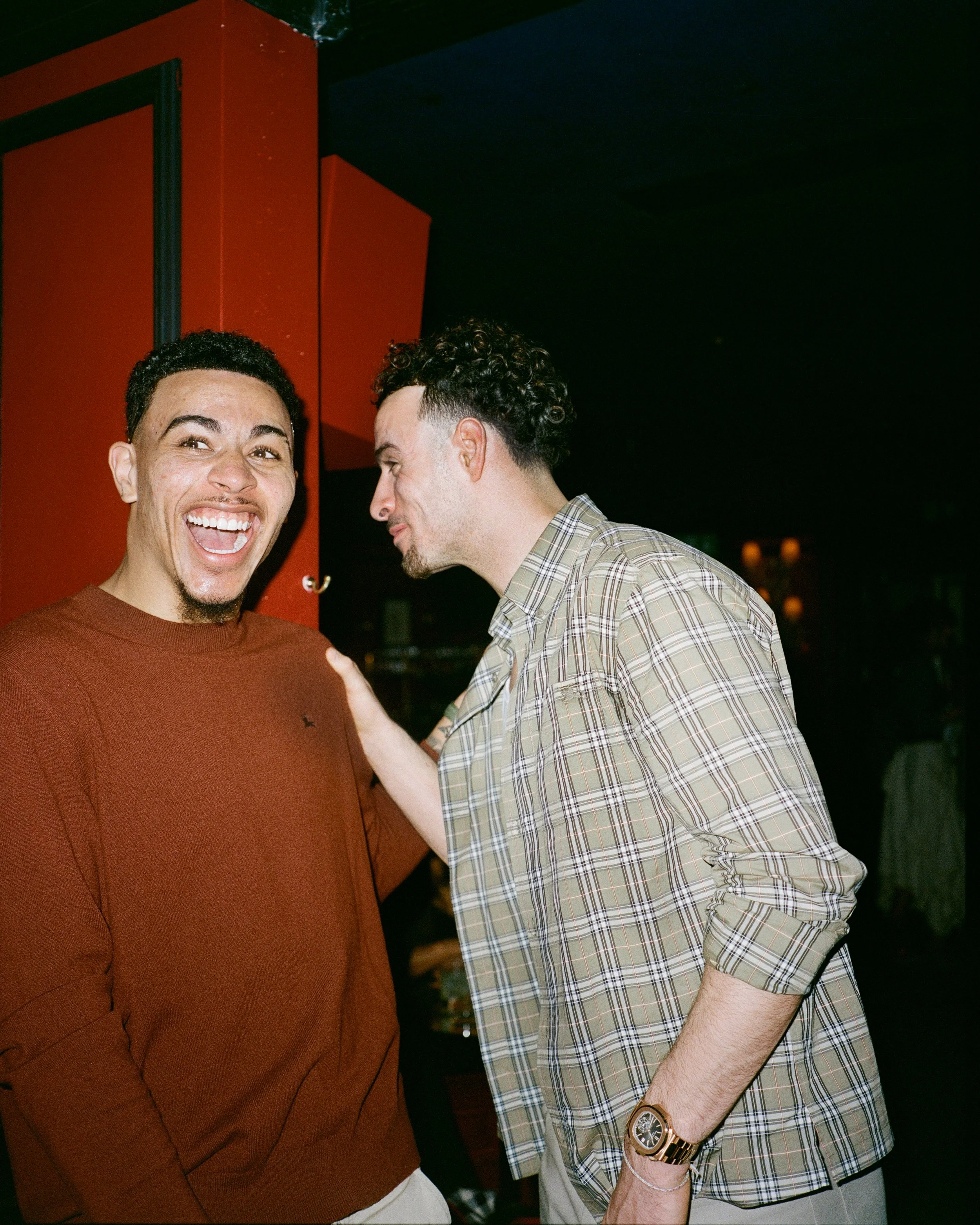Two men are standing close together, one smiling widely and the other looking down and laughing, in a dimly lit indoor setting.