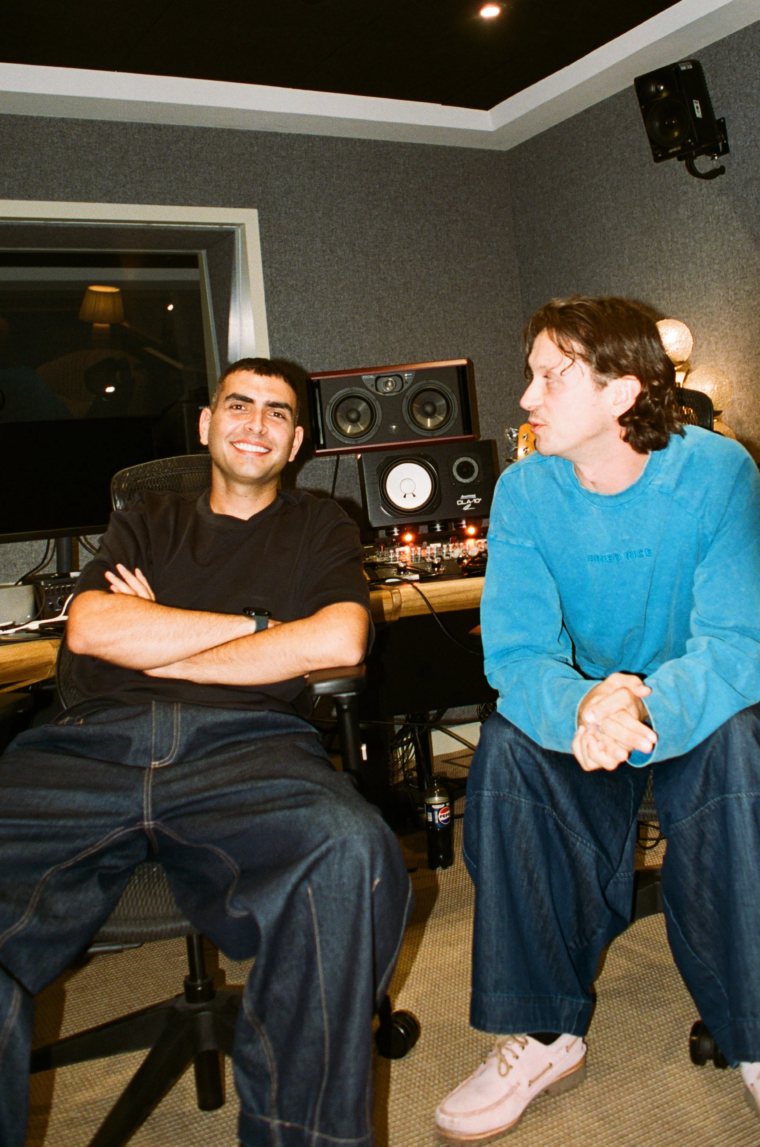 Two men in a recording studio or music production room, one seated with arms crossed and smiling, the other standing and talking. There are studio monitors, audio equipment, and soundproofing on the walls.